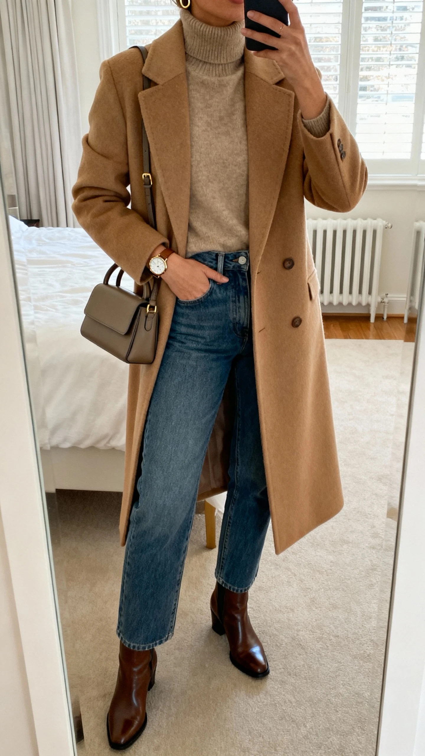 iPhone mirror selfie of a woman in a camel tailored wool coat over a cashmere turtleneck, high-waisted straight-leg jeans, polished leather ankle boots, structured top-handle bag, minimal gold hoops and classic watch; face not visible, cropped at neck, bedroom mirror, natural window light, natural iPhone photo quality.