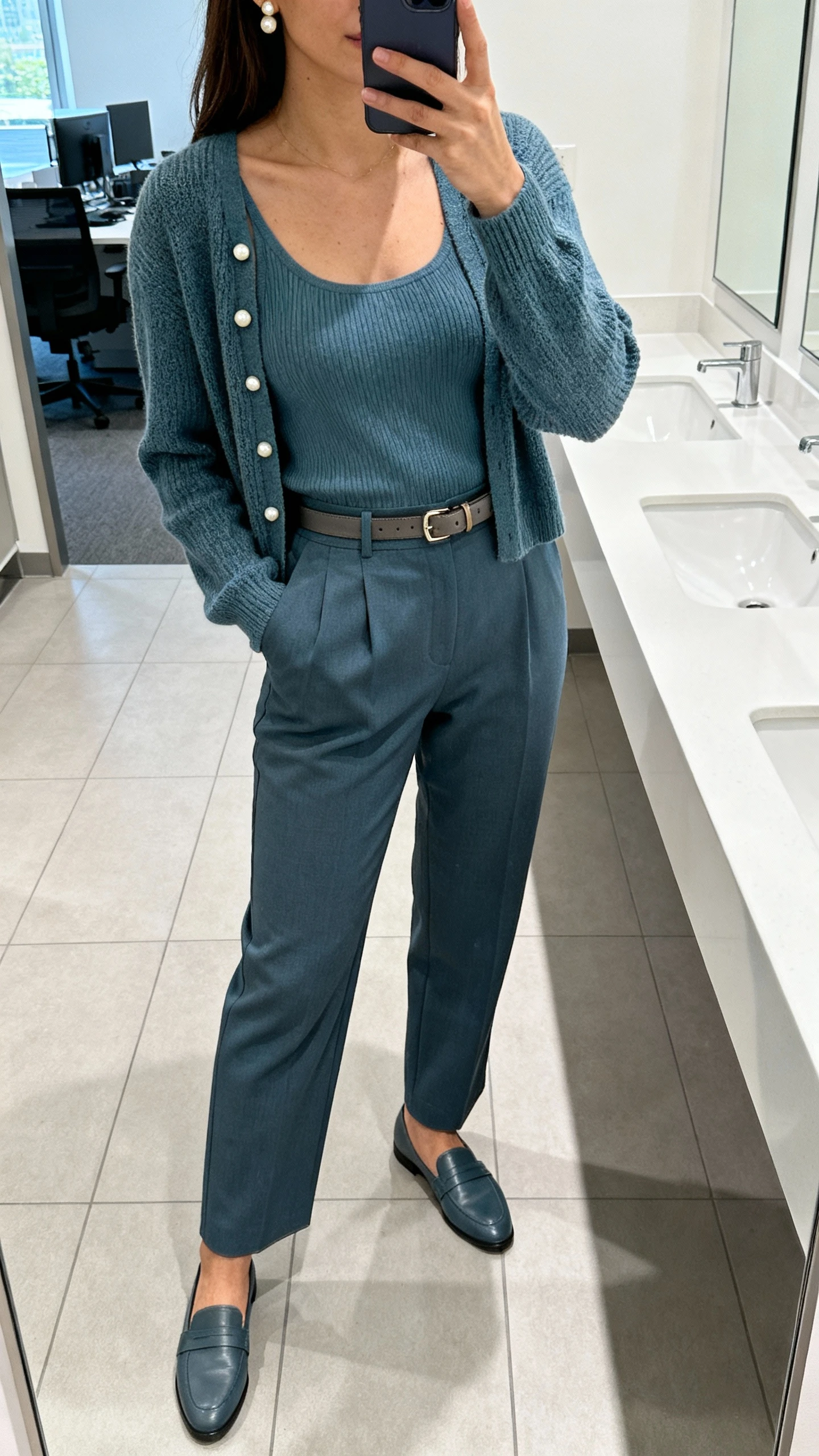 iPhone mirror selfie of a woman in a cardigan set (fine-knit cardigan plus matching knit tank in dusty blue) with high-waist tailored trousers, loafers, pearl studs, slim leather belt, face not visible, office restroom mirror, bright indoor lighting, natural iPhone aesthetic.