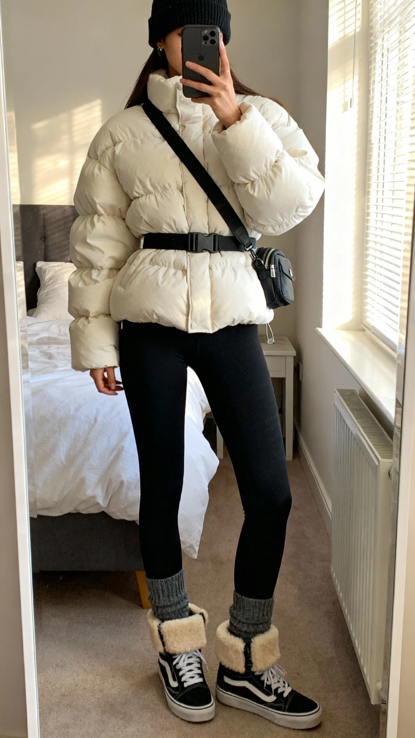 iPhone mirror selfie of a woman in a cloud-puff puffer jacket, black high-waist leggings, chunky socks, shearling-lined sneakers, beanie, crossbody belt bag, face not visible, cropped at neck, bedroom full-length mirror, soft morning window light, natural iPhone photo quality.