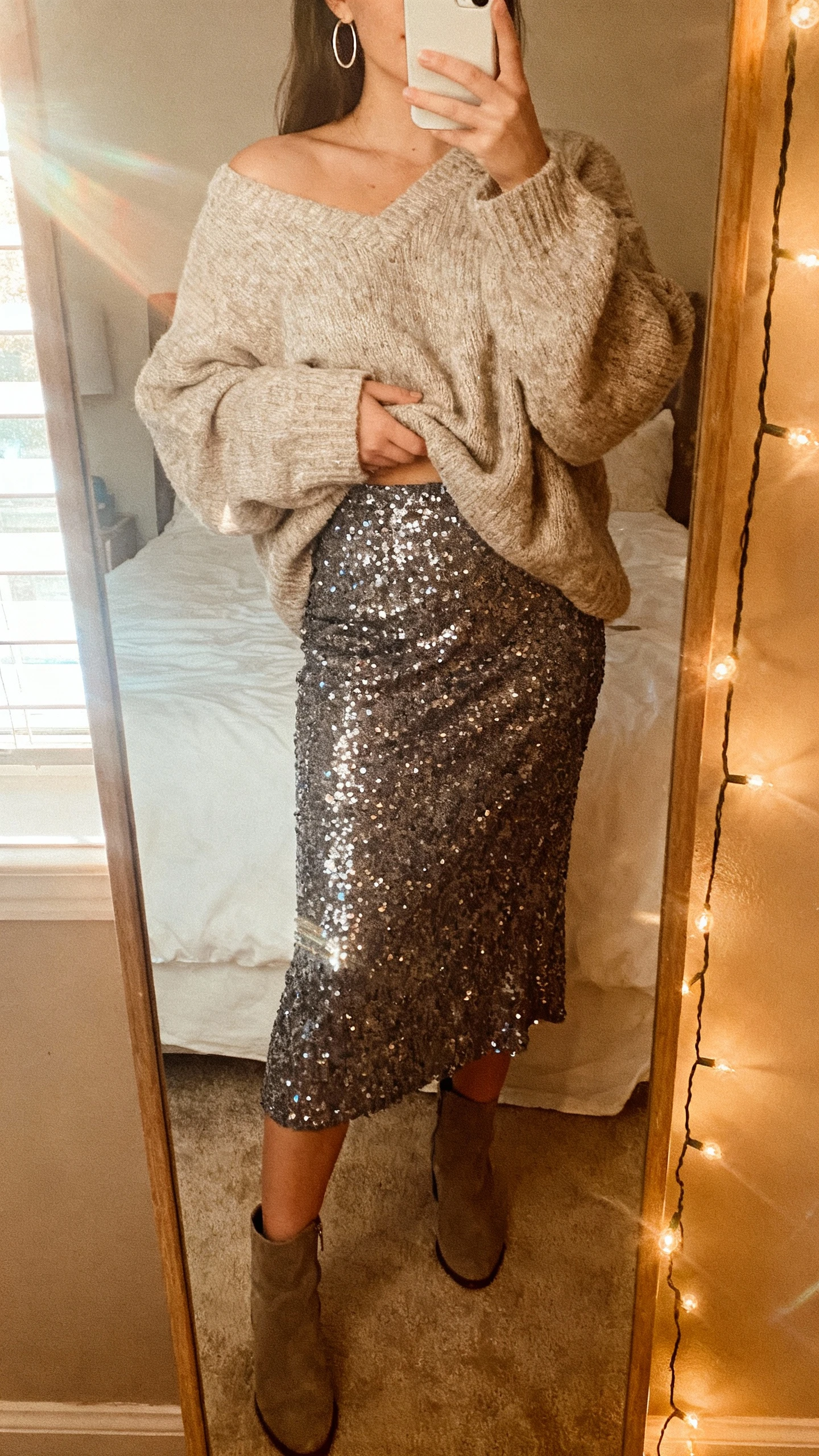 iPhone mirror selfie of a woman in a cozy oversized sweater tucked into a sequin midi skirt, ankle boots, simple hoops, face not visible, bedroom mirror with string lights, natural morning window light, casual iPhone aesthetic.
