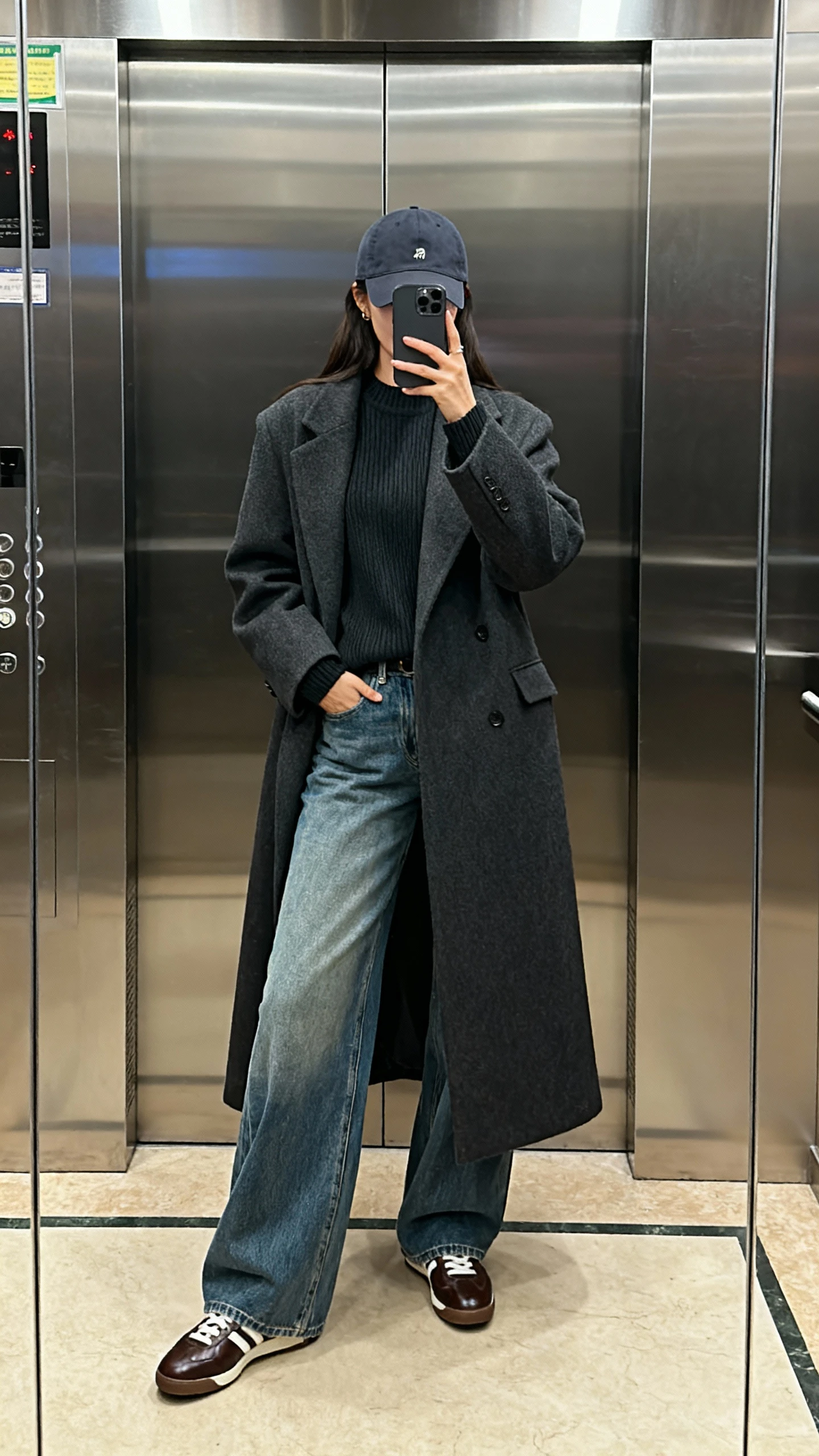 iPhone mirror selfie of a woman in a long maxi coat, wide-leg jeans, sleek sweater, baseball cap, and leather sneakers, face not visible, elevator mirror with indoor lighting, natural iPhone photo quality.
