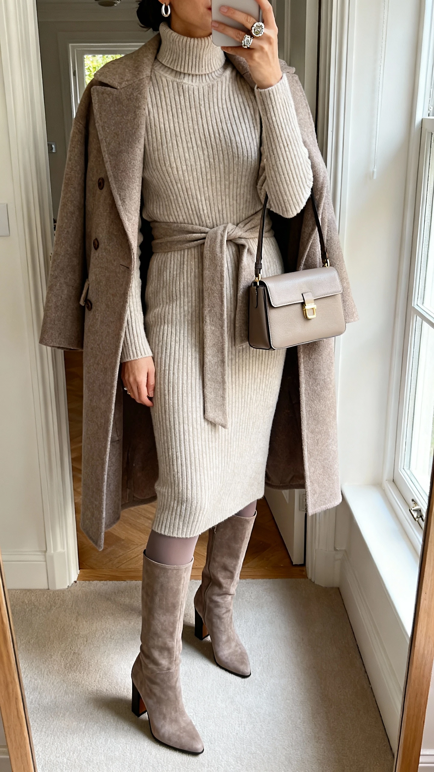 iPhone mirror selfie of a woman in a mid-length ribbed turtleneck sweater dress, knee-high block-heel boots, belted wool wrap coat, opaque tights, structured mini tote, statement ring and minimalist earrings; face not visible, cropped at shoulders, entryway mirror, afternoon window light.