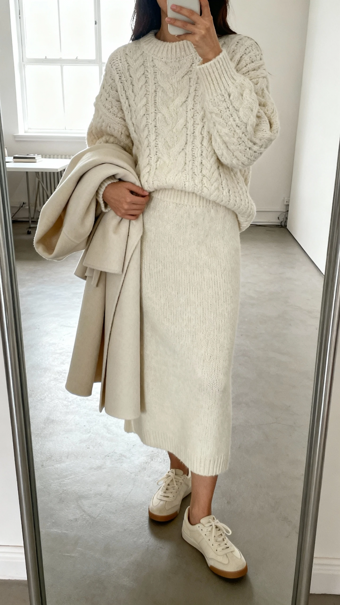 iPhone mirror selfie of a woman in a monochrome knit set (chunky sweater and matching knit skirt), tonal coat draped over arm, minimalist sneakers, face not visible, studio apartment mirror with bright window light, natural iPhone photo quality.