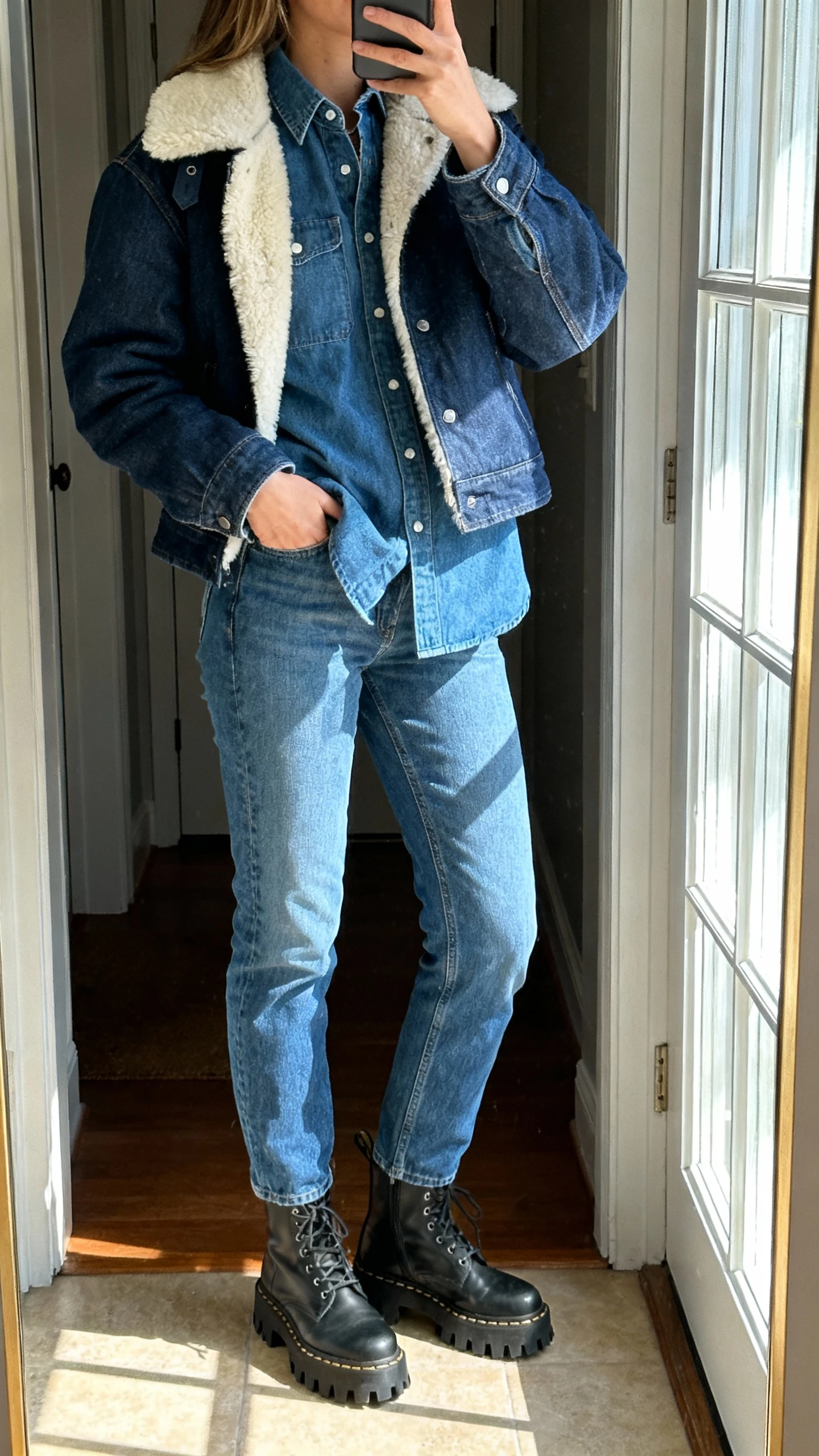 iPhone mirror selfie of a woman in double denim (jeans and denim shirt/jacket) with a shearling-lined jacket layered on top, lug-sole boots, face not visible, entryway mirror with window light, natural iPhone photo quality.