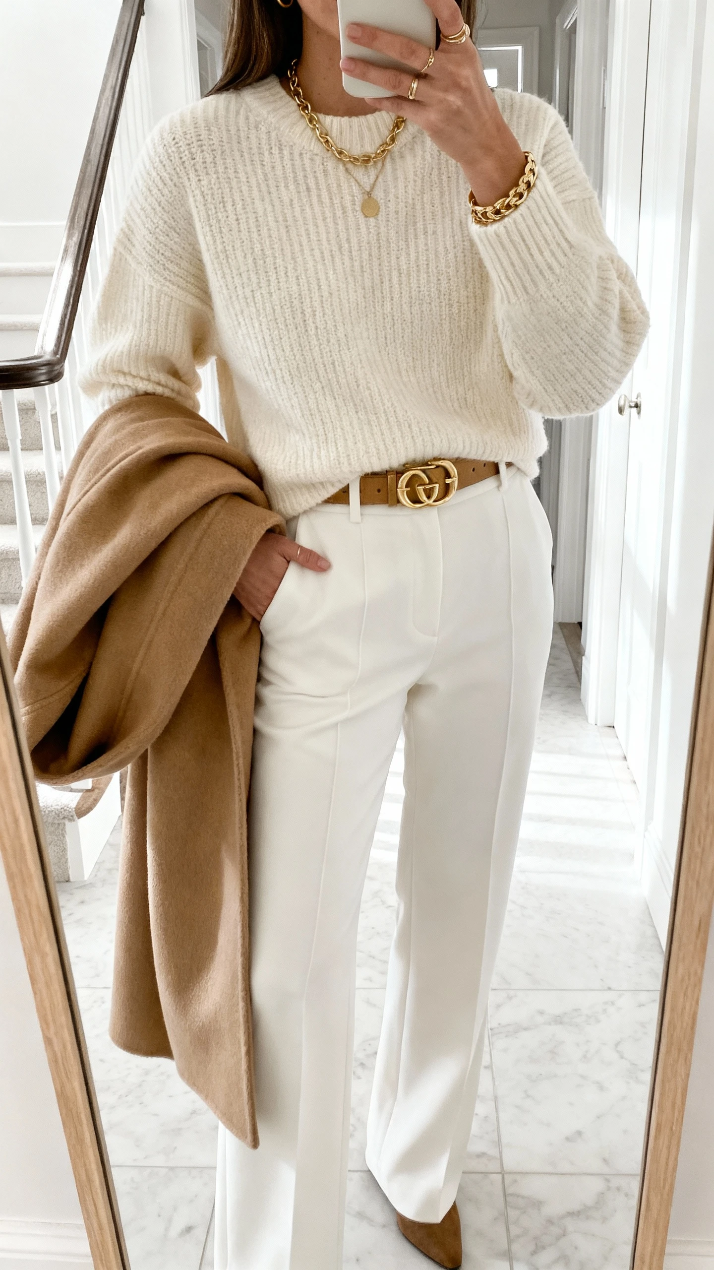 iPhone mirror selfie of a woman in winter white monochrome: cream sweater, white tailored trousers, gold belt and jewelry, camel coat draped over arm, face not visible, entryway mirror, bright natural light, casual iPhone photo.