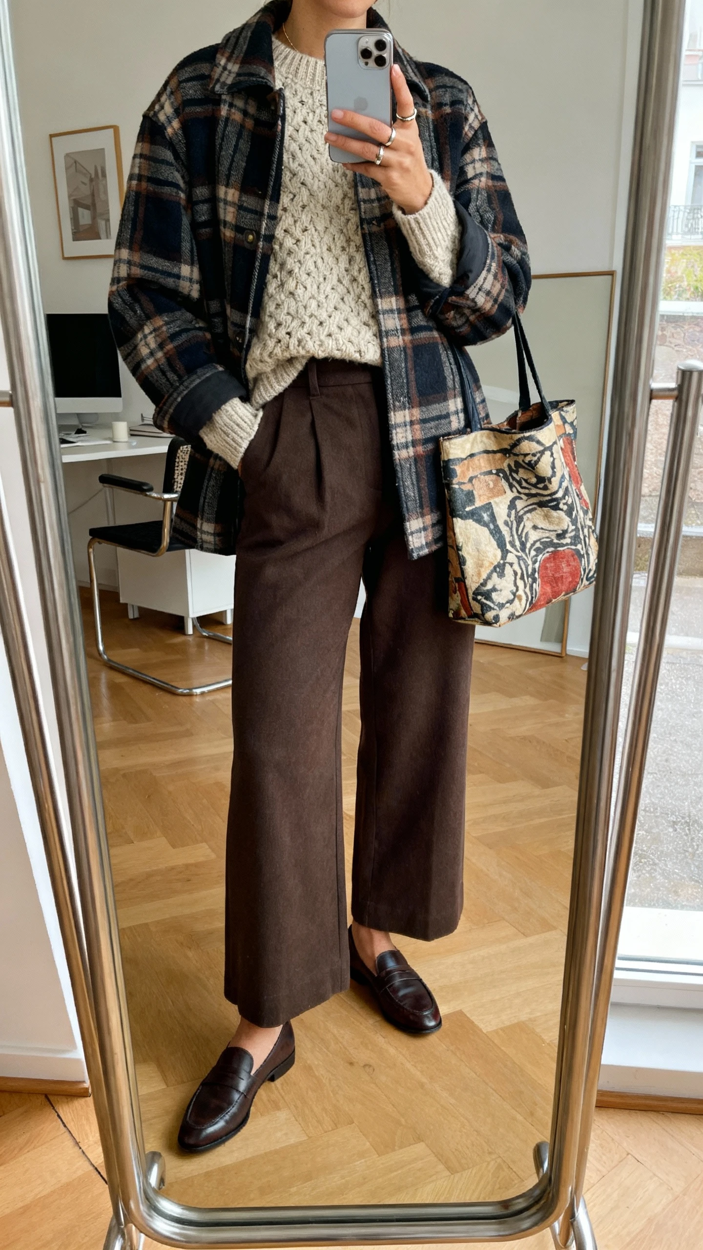 iPhone mirror selfie of a woman layering a plaid shacket over a textured knit sweater with high-waisted wide-leg trousers, loafers, artsy tote, minimal rings, face not visible, studio apartment mirror with diffused daylight, natural iPhone photo