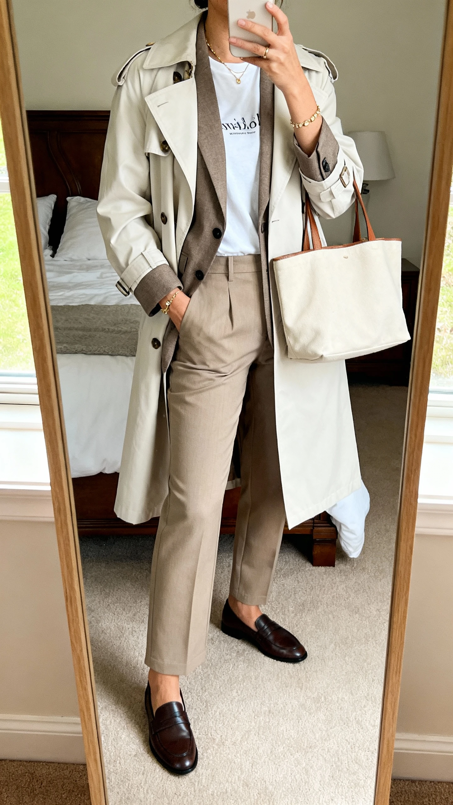 iPhone mirror selfie of a woman summarizing the layered chic: light trench over a blazer and tee with straight trousers, loafers, subtle jewelry, tote bag, face not visible, bedroom mirror, soft morning window light, natural iPhone photo quality.