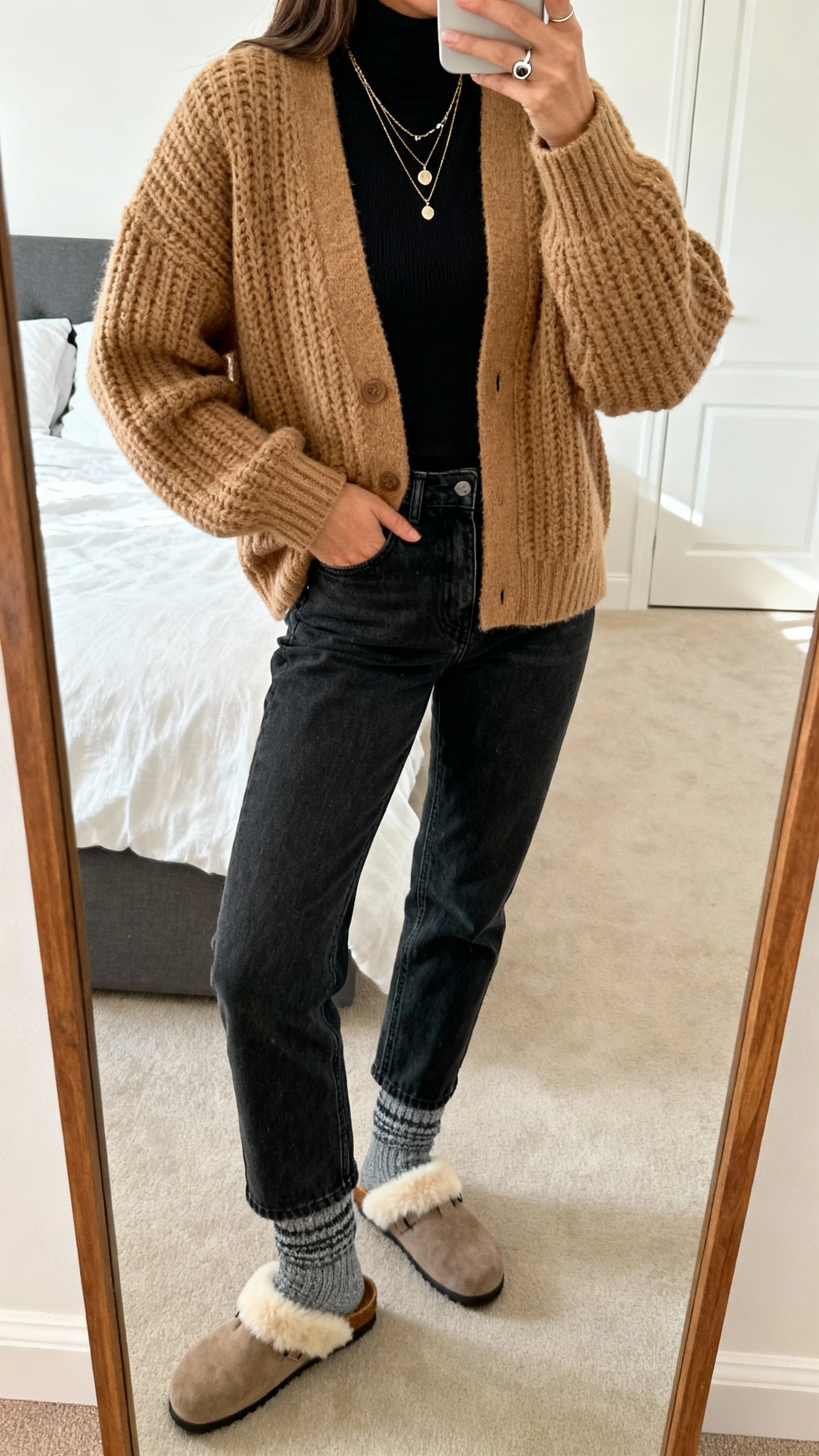 iPhone mirror selfie of a woman wearing a chunky tan cardigan over a fitted black turtleneck, dark-wash straight-leg jeans, shearling-lined clogs, cozy marled gray socks, layered necklaces and a signet ring, face not visible, cropped at shoulders, bedroom mirror, natural daylight, iPhone photo quality.