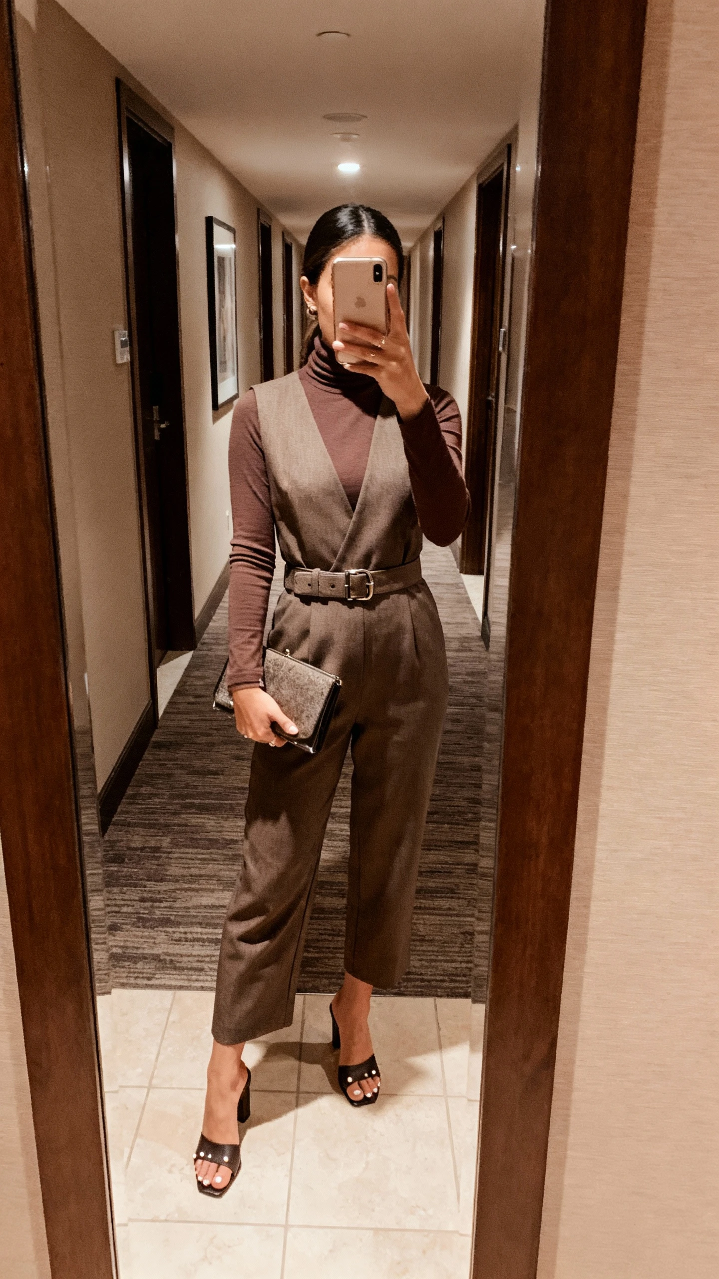 iPhone mirror selfie of a woman wearing a fitted turtleneck under a tailored dressy jumpsuit, waist belt, heeled mules, simple studs, clutch in hand, face not visible, hotel hallway mirror, evening indoor lighting, natural iPhone photo.