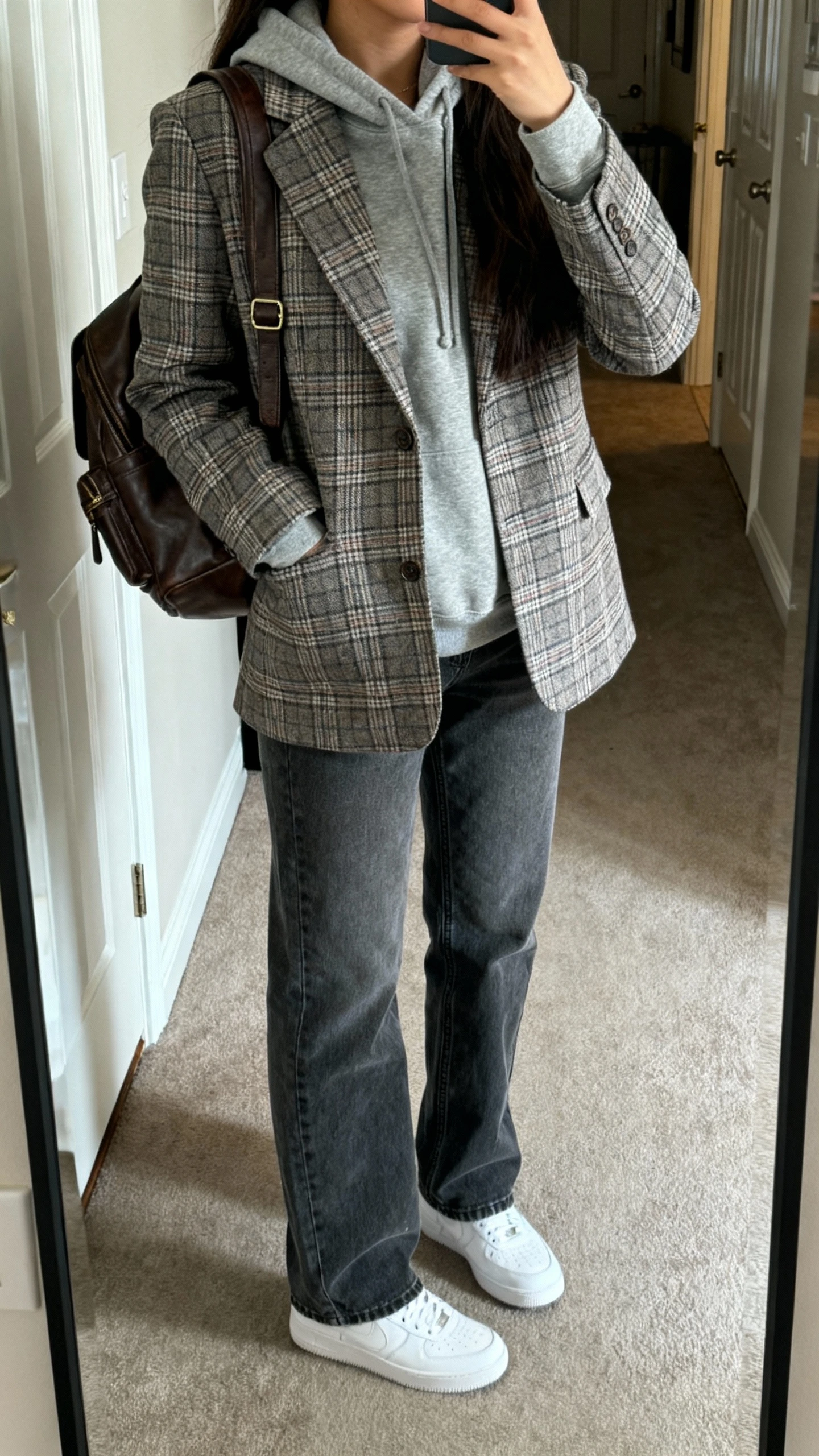 iPhone mirror selfie of a woman wearing a gray hoodie layered under a plaid blazer with straight-leg jeans, white sneakers, and a leather backpack, face not visible, cropped at shoulders, apartment entryway mirror, natural daylight, casual iPhone photo.