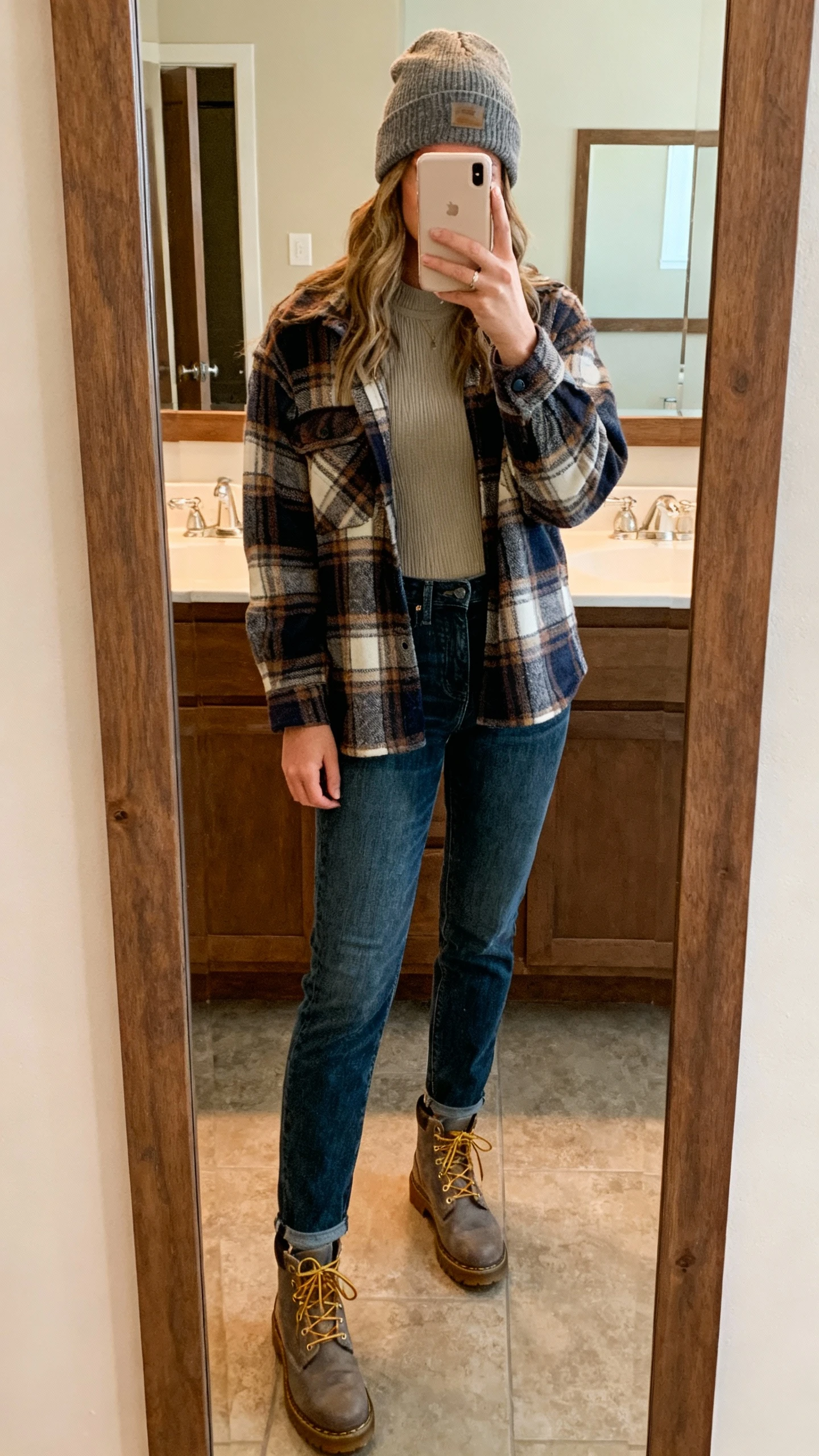 iPhone mirror selfie of a woman wearing a plaid shacket over thermal layers, straight jeans, beanie, and lace-up boots, face not visible, bathroom mirror with warm indoor light, natural iPhone photo quality.
