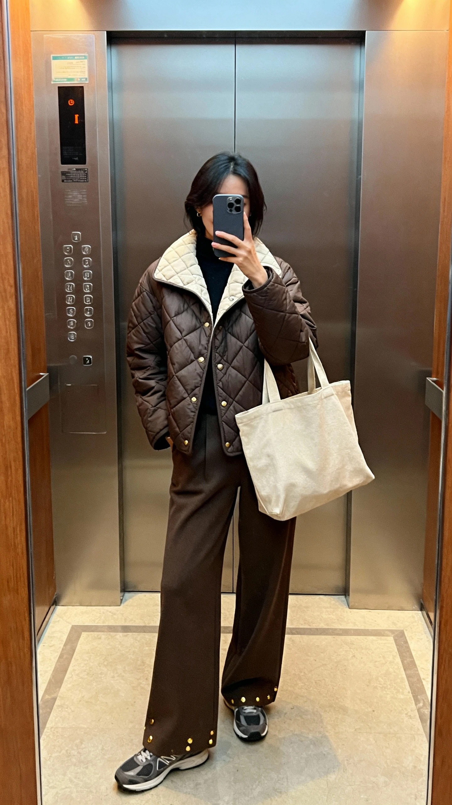 iPhone mirror selfie of a woman wearing a quilted liner jacket with wide-leg trousers, sleek sneakers, minimal gold studs, and a canvas tote, face not visible, elevator mirror, indoor lighting, natural iPhone photo.