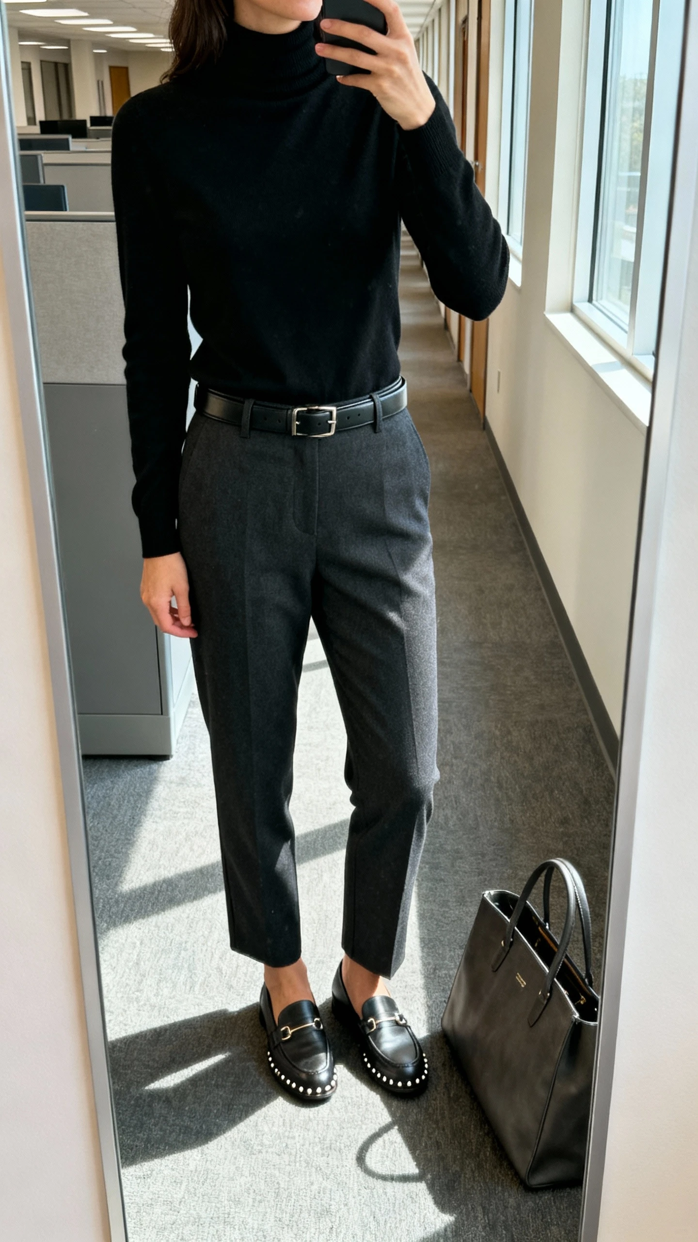 iPhone mirror selfie of a woman wearing a sleek black turtleneck tucked into tailored charcoal trousers with a slim leather belt, loafers, simple studs, and a structured tote on the floor, face not visible, cropped at neck, office hallway mirror, morning window light, natural iPhone photo quality.