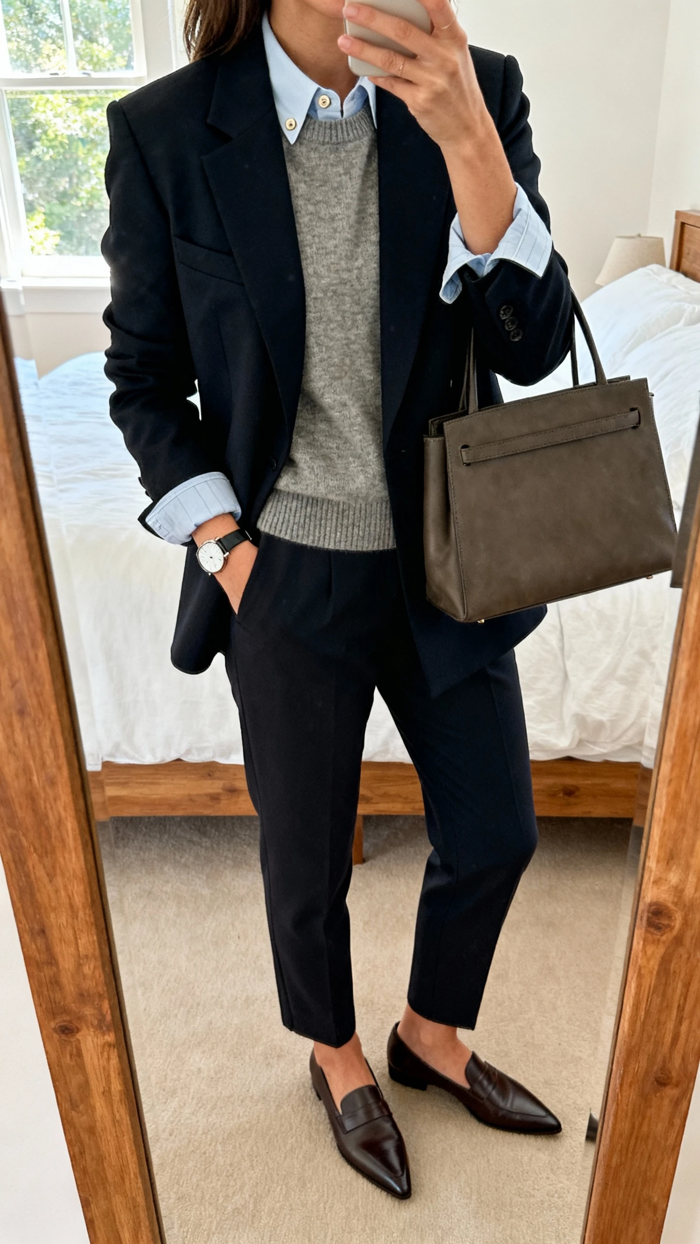 iPhone mirror selfie of a woman wearing a tailored blazer layered over a fine knit sweater with a crisp button-down peeking at collar and cuffs, tailored trousers, pointed loafers, simple watch and structured tote, face not visible, cropped at neck, bedroom mirror, natural window light, natural iPhone photo quality.