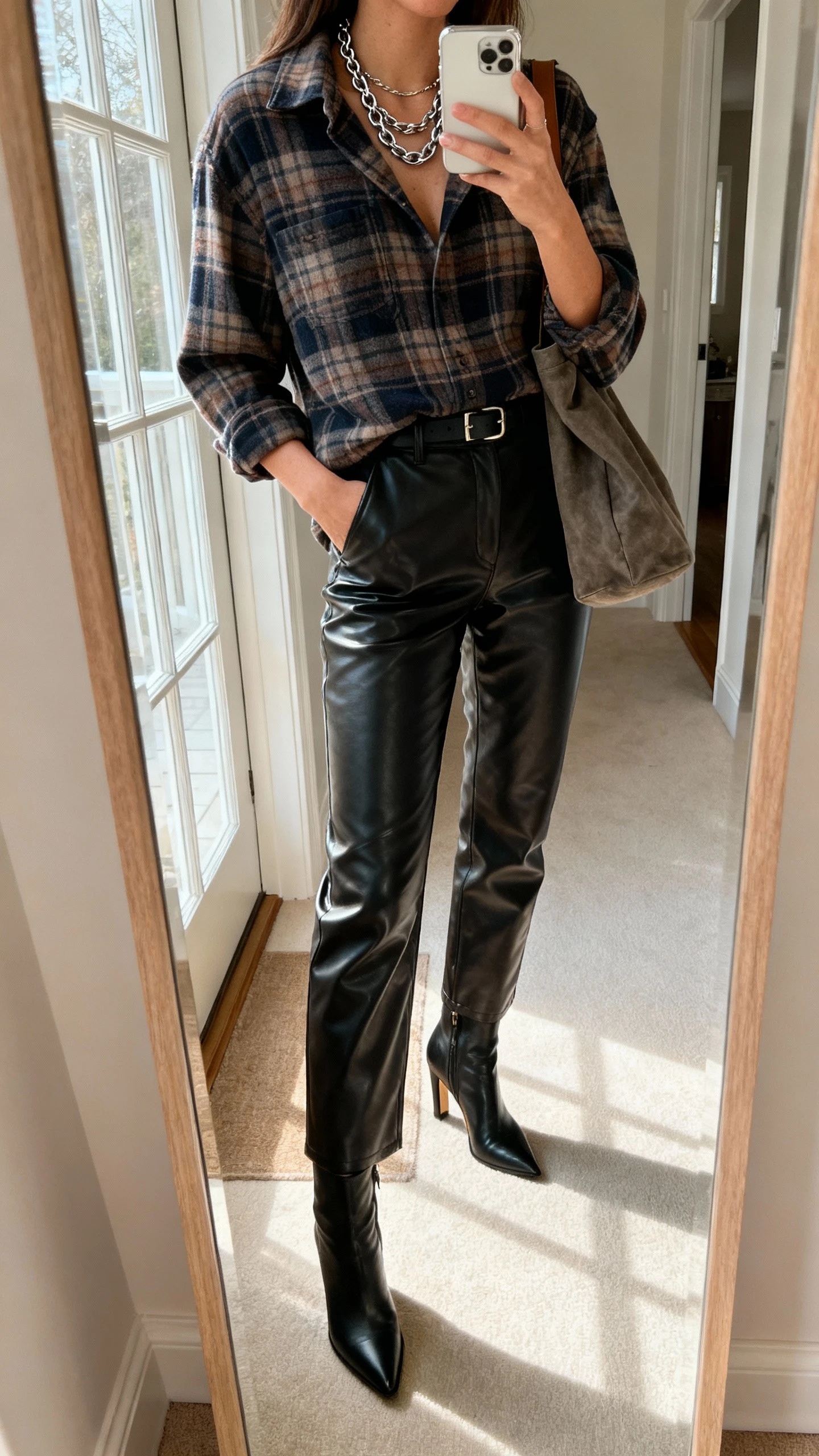 iPhone mirror selfie of a woman wearing an elevated flannel shirt tucked into faux leather pants, heeled ankle boots, layered chain necklace, slouchy shoulder bag, face not visible, entryway mirror, morning window light, natural iPhone quality.