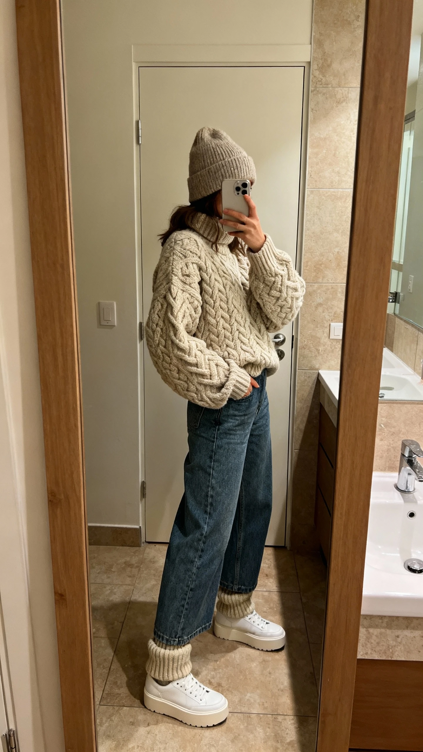 iPhone mirror selfie of a woman wearing wide-leg jeans, a chunky cable-knit sweater, white platform sneakers, wool beanie, and thick crew socks peeking, face not visible, side angle in bathroom mirror, warm indoor lighting, iPhone photo quality.