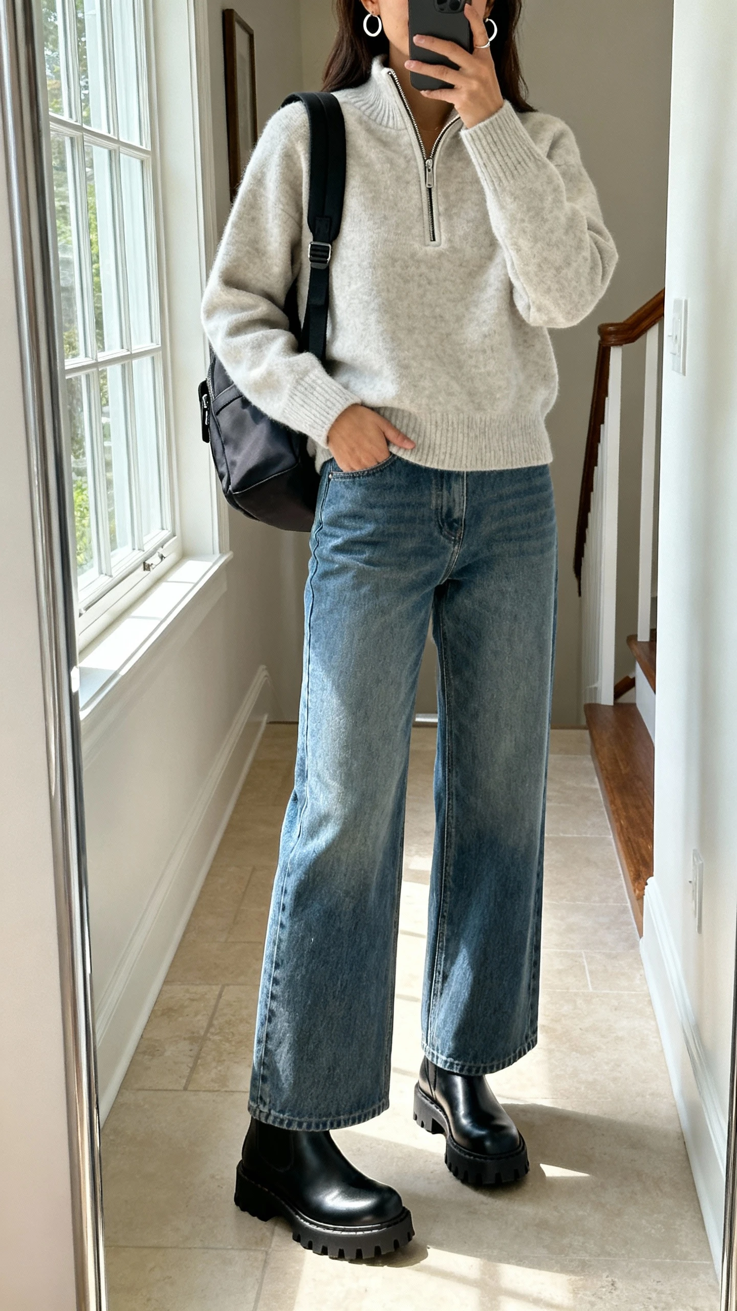 iPhone mirror selfie of a woman wearing wide-leg jeans, a half-zip sweater, black lug-sole Chelsea boots, minimal hoop earrings, and a structured backpack, face not visible, cropped at shoulders, entryway mirror, bright window light, iPhone photo quality.