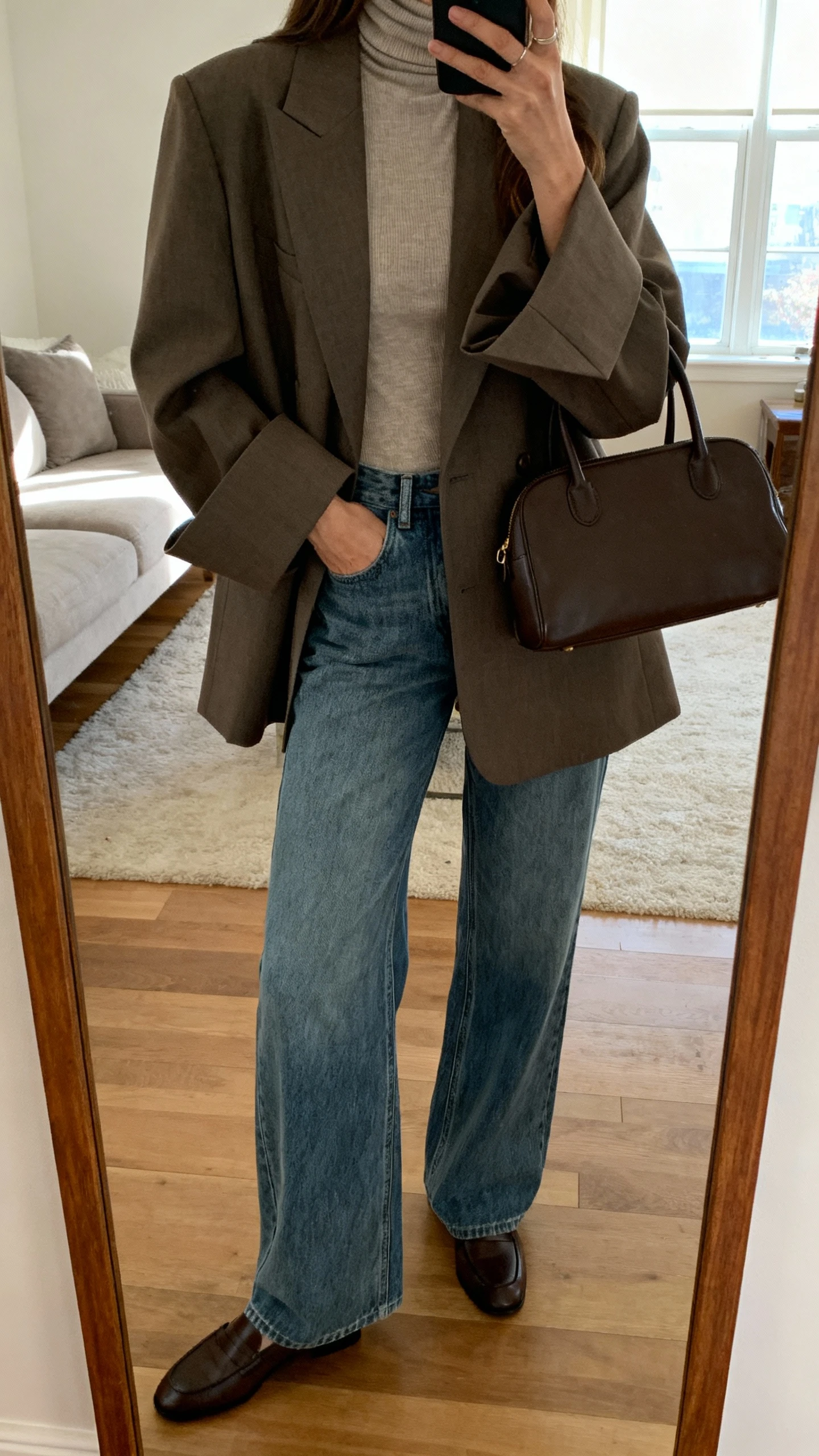 iPhone mirror selfie of a woman wearing wide-leg jeans, a fitted turtleneck under an oversized blazer with dramatic sleeves, loafers, and a top-handle bag, face not visible, cropped at shoulders, living room full-length mirror, natural window light, iPhone photo quality.
