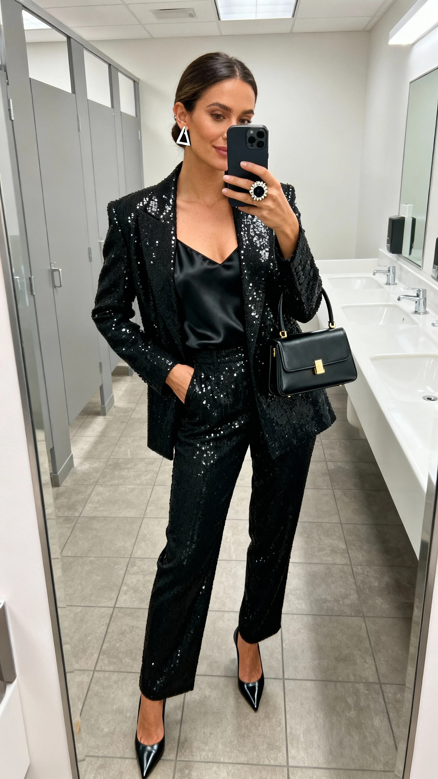 iPhone office restroom mirror selfie of a woman in a black sequined blazer with matching trousers, black satin camisole, pointed-toe pumps, structured mini top-handle bag, geometric stud earrings, and a bold cocktail ring; face not visible, bright indoor lighting, natural iPhone aesthetic.