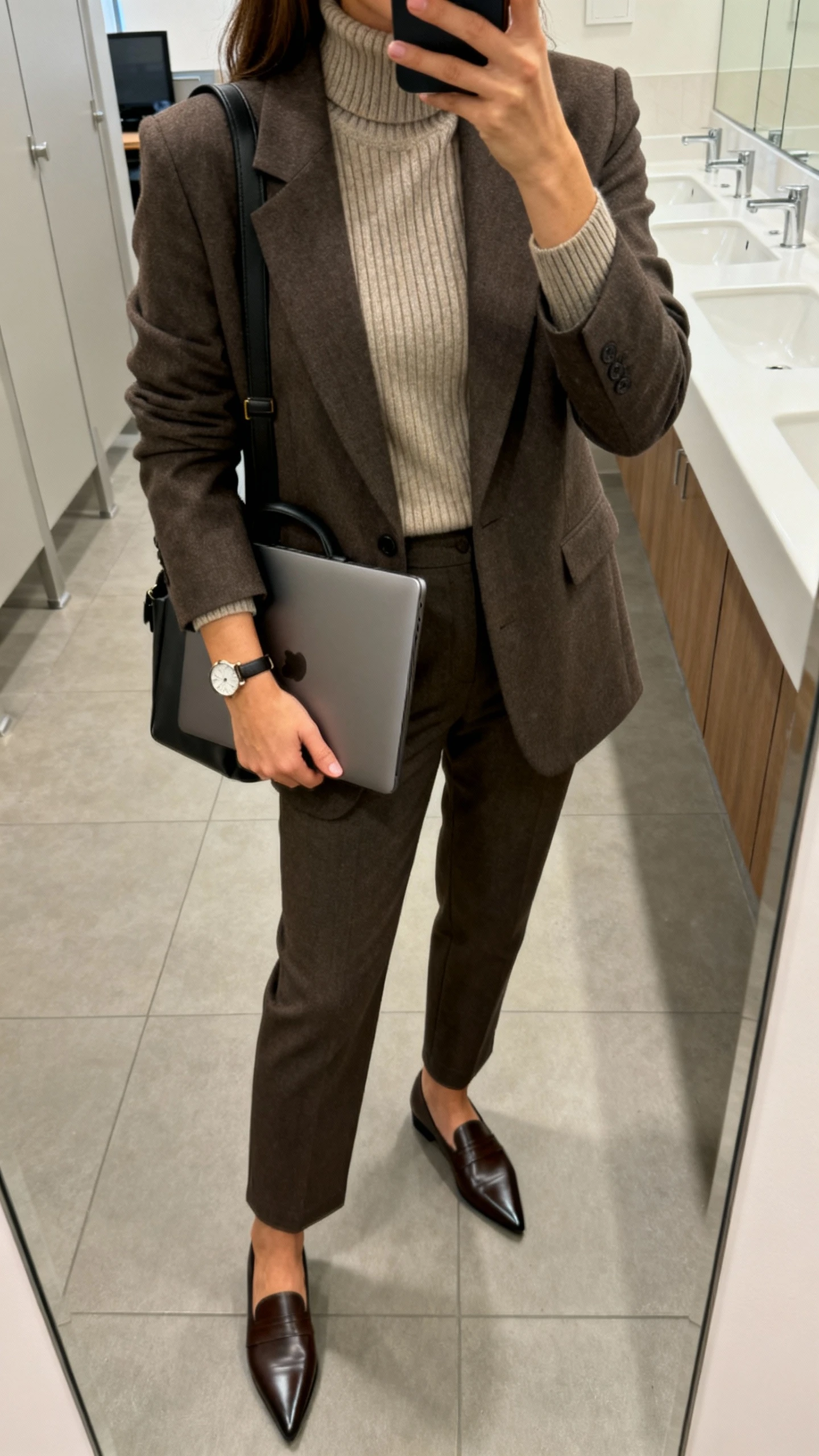 iPhone office restroom mirror selfie of a woman in a tailored blazer over a cozy ribbed turtleneck, straight-leg trousers, pointed-toe loafers, simple watch, structured laptop bag, face not visible, overhead office lighting, natural iPhone aesthetic.