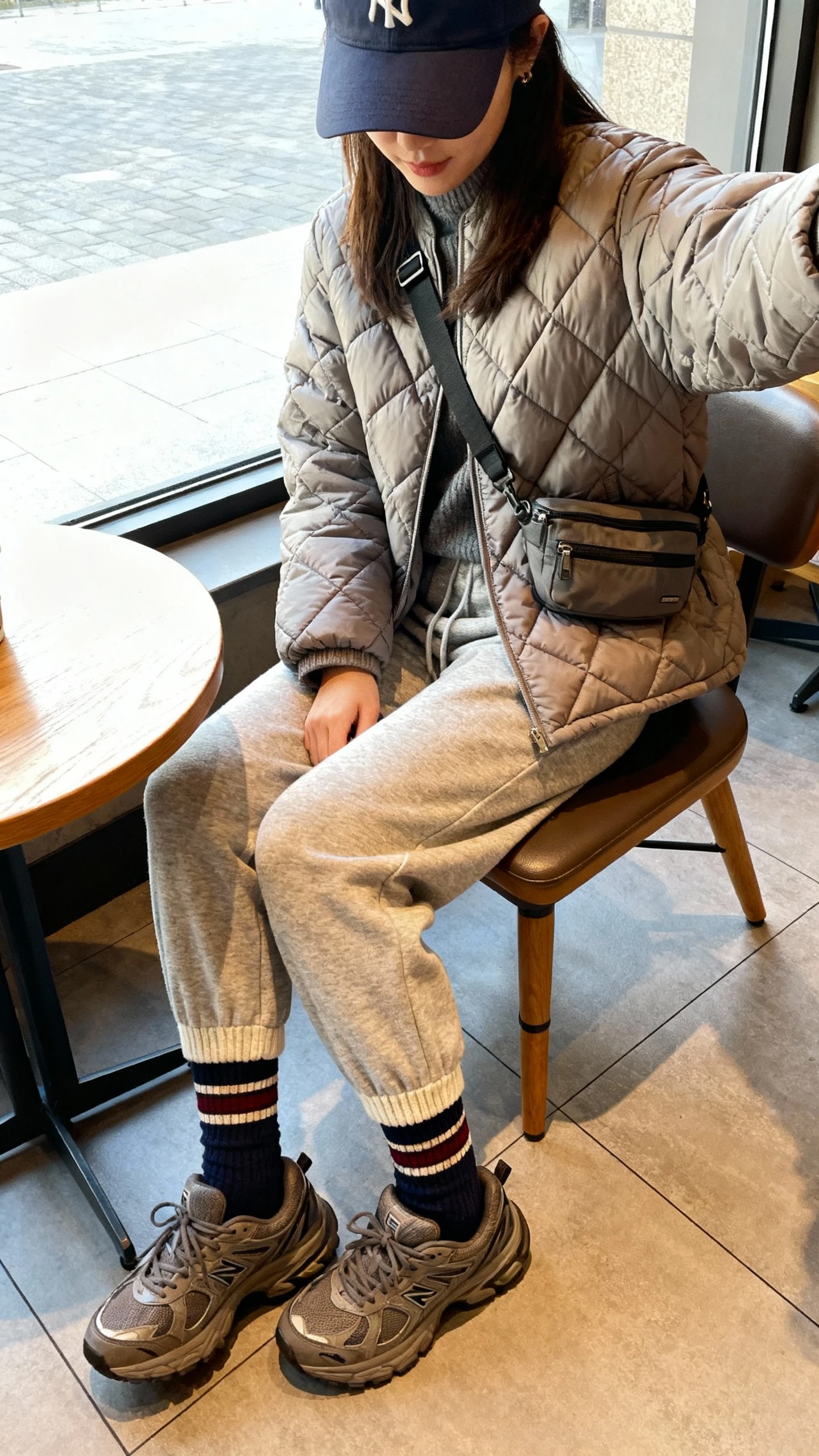 iPhone seated cafe selfie cropped at shoulders of a woman in a quilted puffer jacket, knit joggers, chunky sneakers, sporty crew socks, baseball cap, crossbody belt bag, face not visible, bright window light, casual iPhone quality