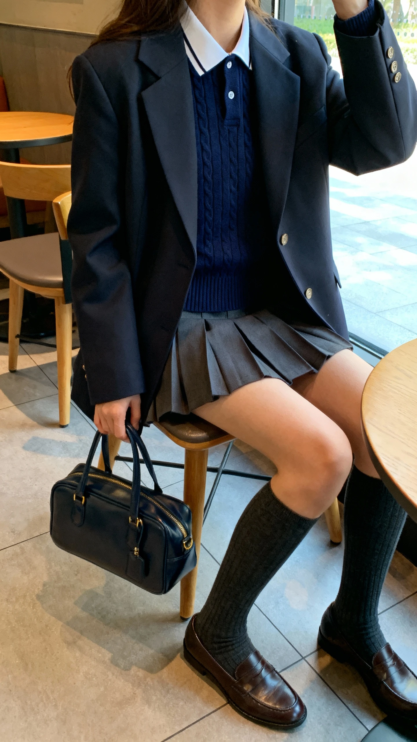 iPhone seated cafe selfie (side angle) of a woman wearing a preppy polo knit sweater (navy with collar) under a tailored blazer, pleated mini skirt, knee socks, loafers, structured satchel, face not visible, window light, casual iPhone aesthetic.