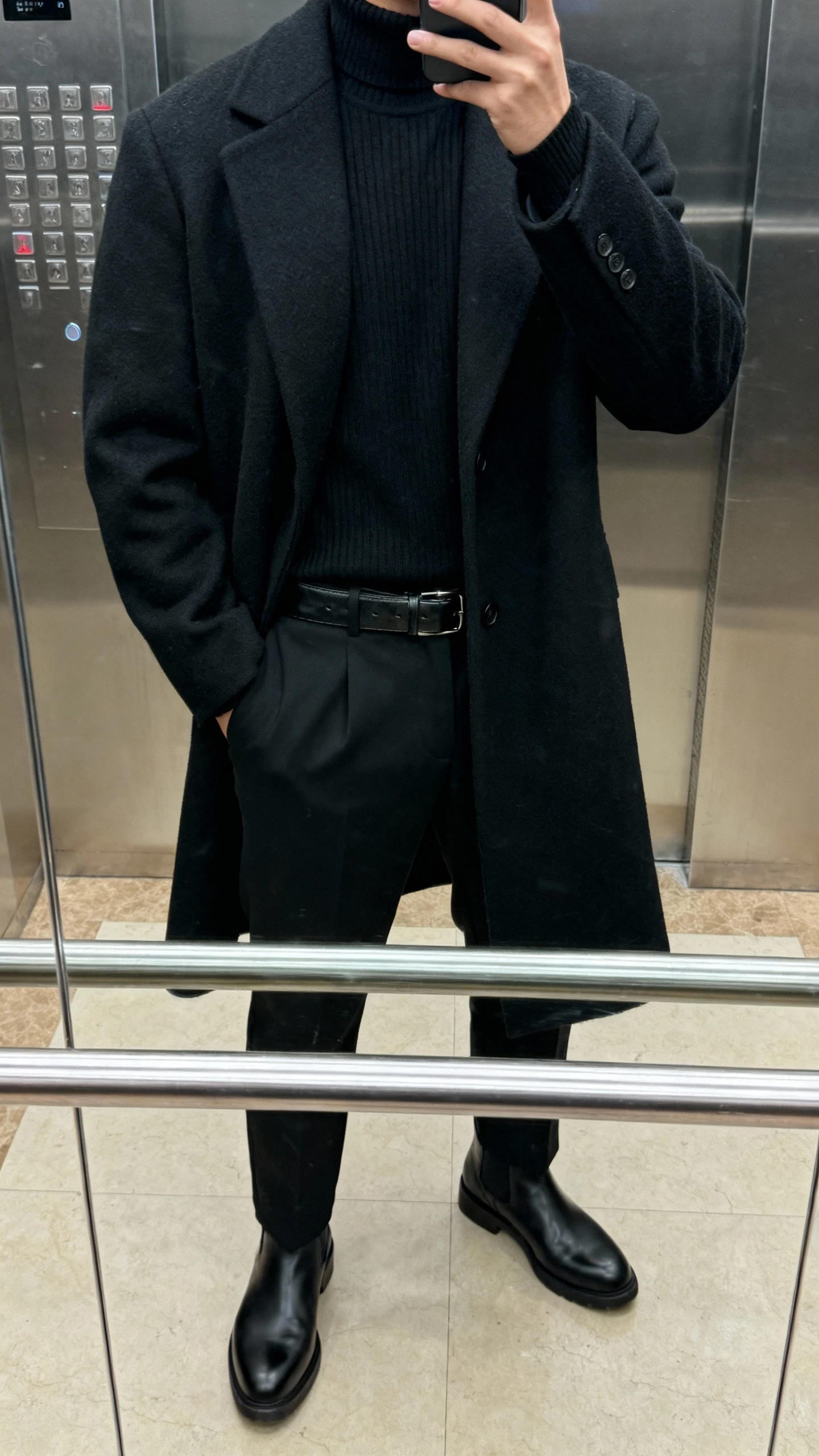 iPhone selfie cropped at shoulders of a man wearing an all-black monochrome fit: black wool overcoat, black mock-neck sweater, black tailored trousers, and black Chelsea boots with a leather belt, taken in an elevator mirror with soft indoor lighting, face not visible, natural iPhone photo quality.
