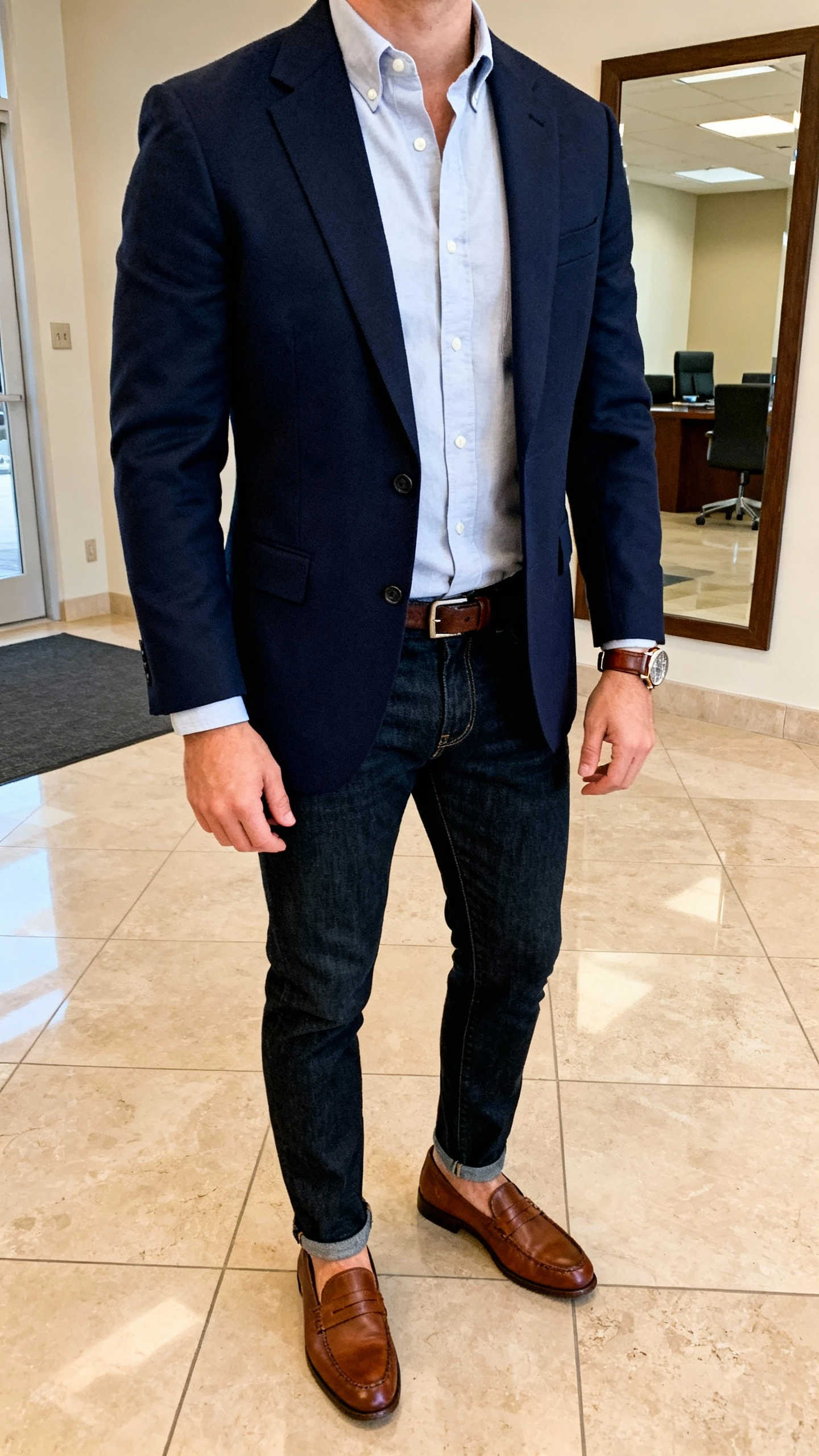 iPhone selfie cropped at shoulders showing a man in a navy blazer over a light Oxford shirt, dark denim jeans, brown loafers, and a leather watch, face not visible, office lobby mirror, warm indoor lighting, natural iPhone aesthetic.