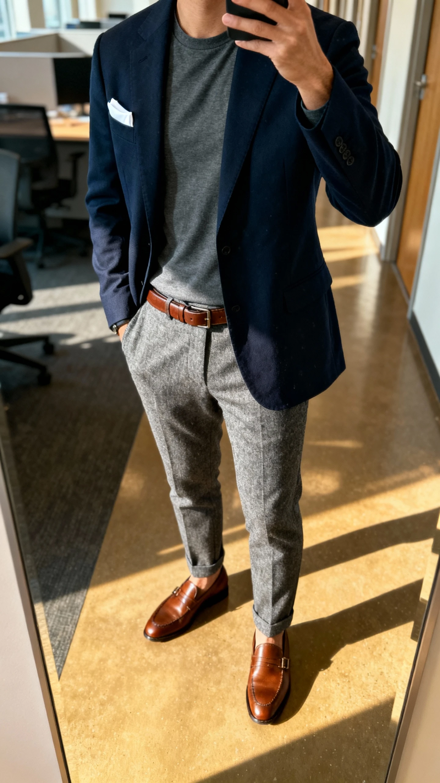 iPhone selfie from chest-down of a man in an unstructured navy blazer over a slate crewneck T-shirt, gray wool trousers, brown leather loafers, slim brown belt, and a white pocket square peeking; office hallway mirror, morning light, face not visible, natural iPhone quality.