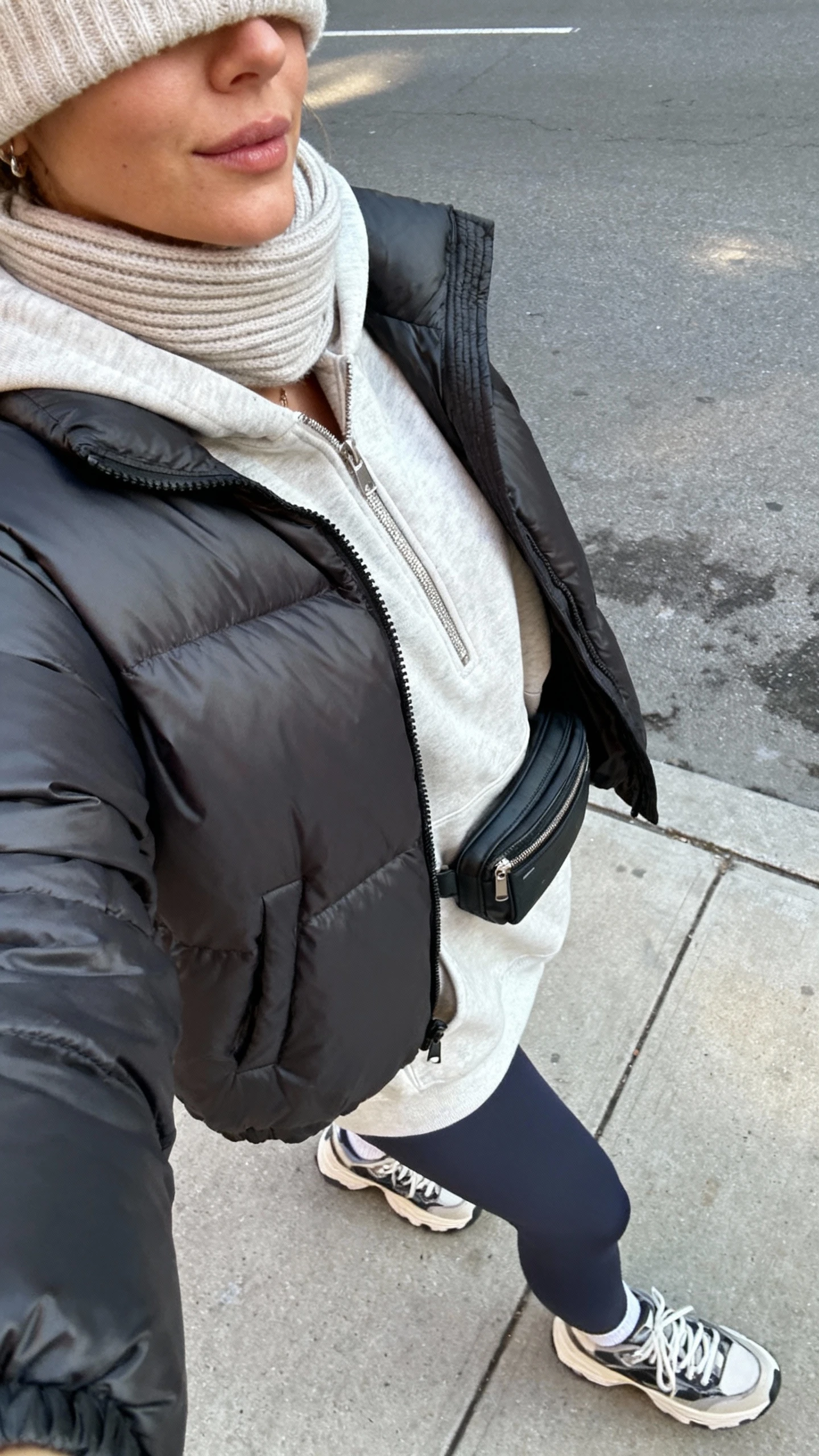 iPhone selfie side angle of a woman in a matte puffer jacket over an oversized zip hoodie, premium leggings, chunky lace-up sneakers, soft beanie and ribbed scarf, compact belt bag; face not visible, taken on a city sidewalk, natural daylight, casual iPhone photo.