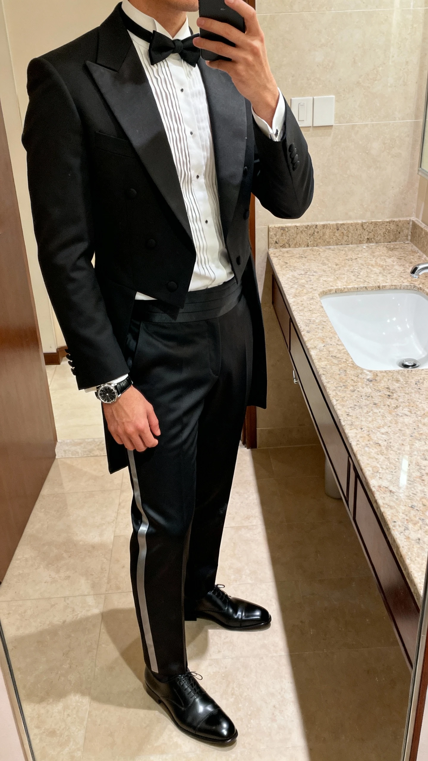 iPhone side-angle bathroom mirror selfie of a man in a modern tuxedo with a peak-lapel jacket, crisp pleated-front shirt, slim black bow tie, satin-trim trousers, polished black oxfords, and a sleek watch, face not visible, warm vanity lighting, natural iPhone photo.