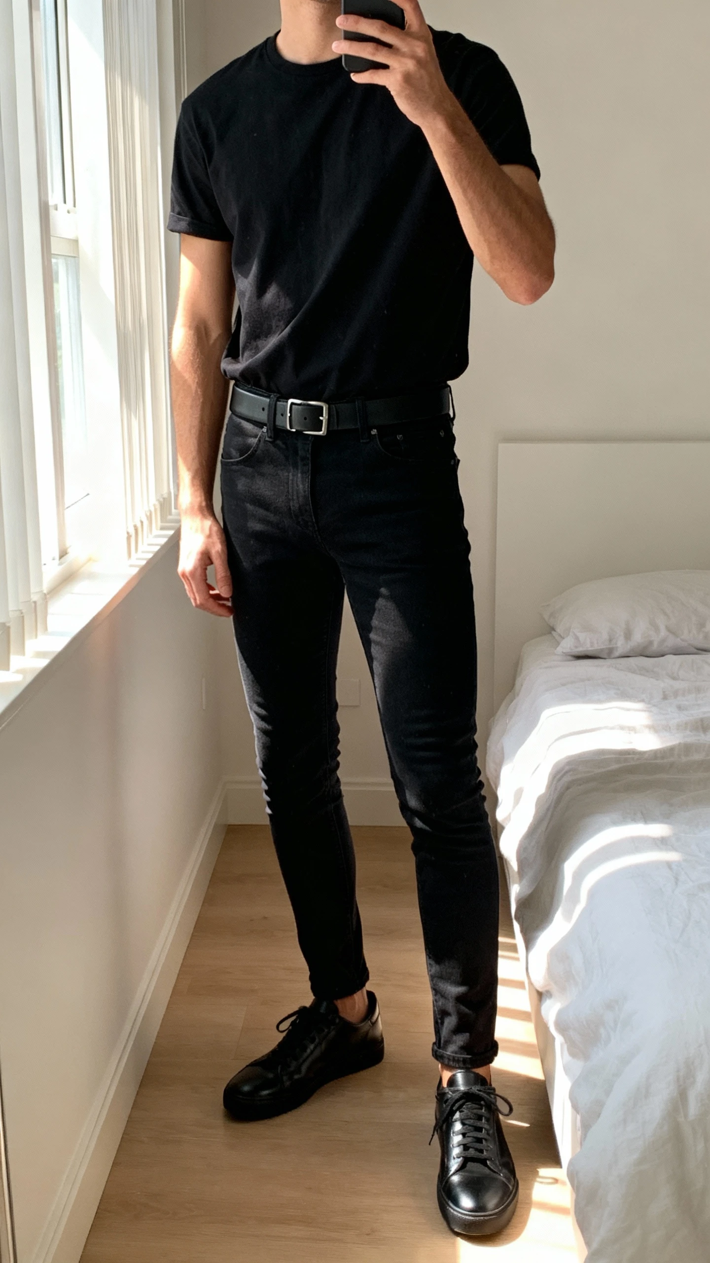 iPhone side-angle bedroom selfie of a man wearing all-black: black tee, black slim jeans, black leather sneakers, and a black belt, face not visible, soft morning window light, natural iPhone photo.