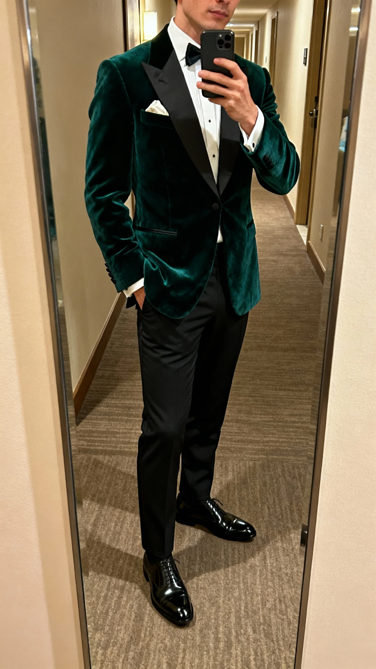 iPhone side-angle mirror selfie of a man in a deep emerald velvet dinner jacket over a crisp white dress shirt, black tuxedo trousers, black patent loafers, pocket square, and a subtle bow tie undone, hotel hallway mirror under warm indoor lighting, face not visible, natural iPhone photo quality.