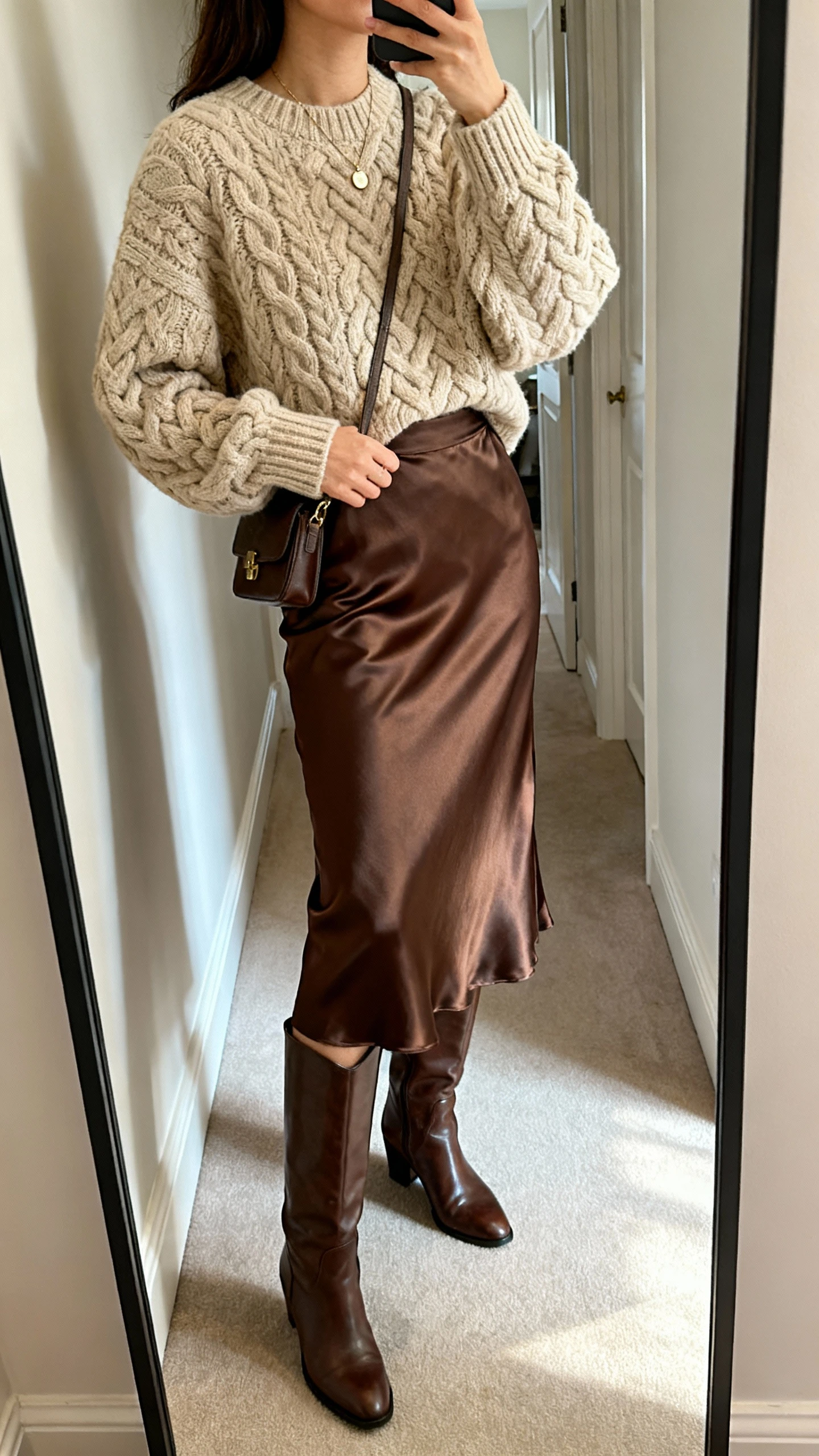 iPhone side-angle mirror selfie of a woman in a chunky cable-knit sweater (oatmeal) tucked into a satin midi skirt (chocolate), knee-high leather boots, delicate pendant necklace, small crossbody bag, face not visible, hallway mirror, soft afternoon light, casual iPhone photo.