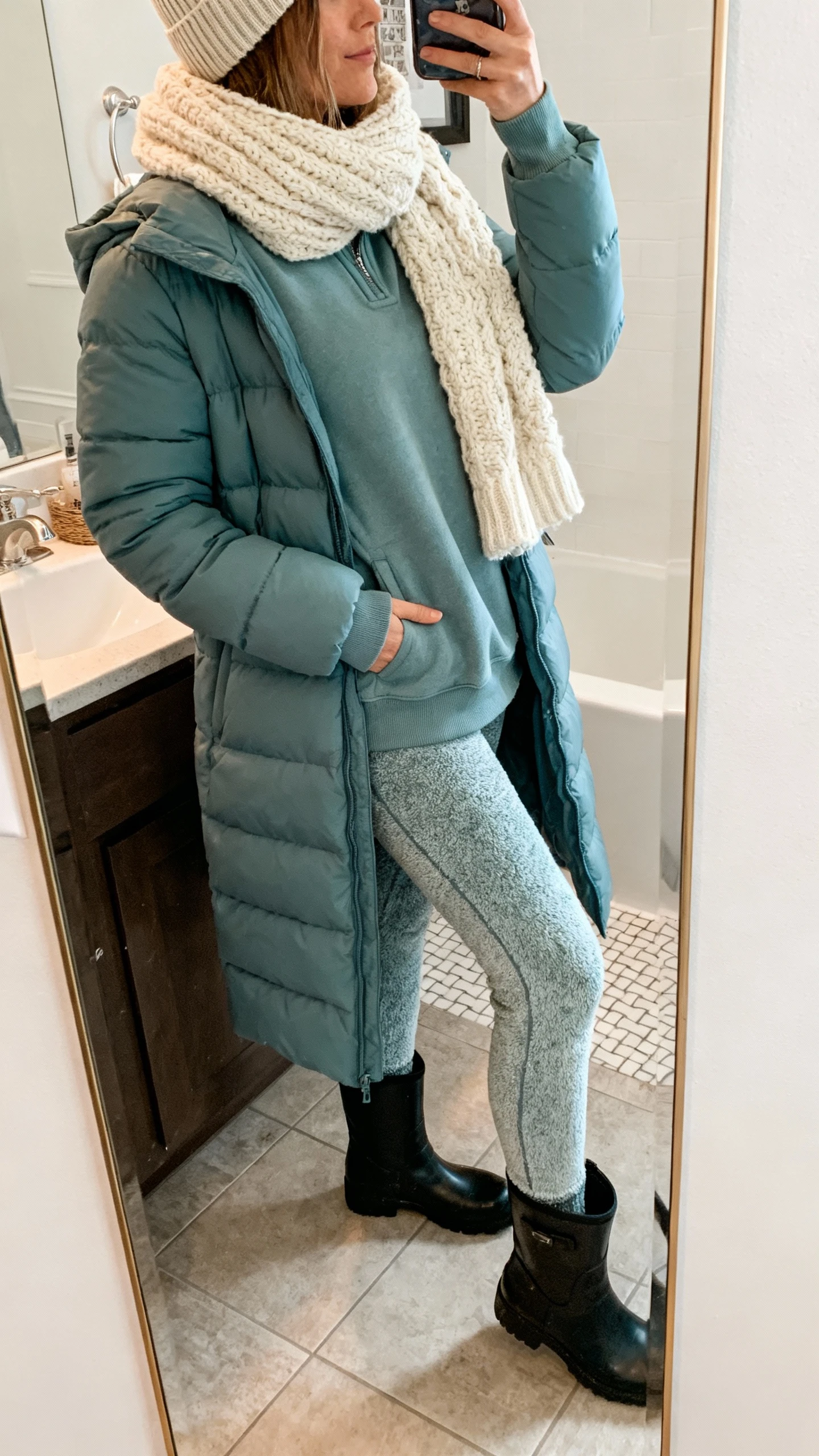 iPhone side-angle mirror selfie of a woman wearing a mid-length puffer in dusty blue, fleece-lined leggings, a cozy mock-neck sweatshirt, chunky cream scarf, waterproof black ankle boots, and a ribbed beanie, face not visible, bathroom mirror, soft indoor lighting, iPhone photo quality.