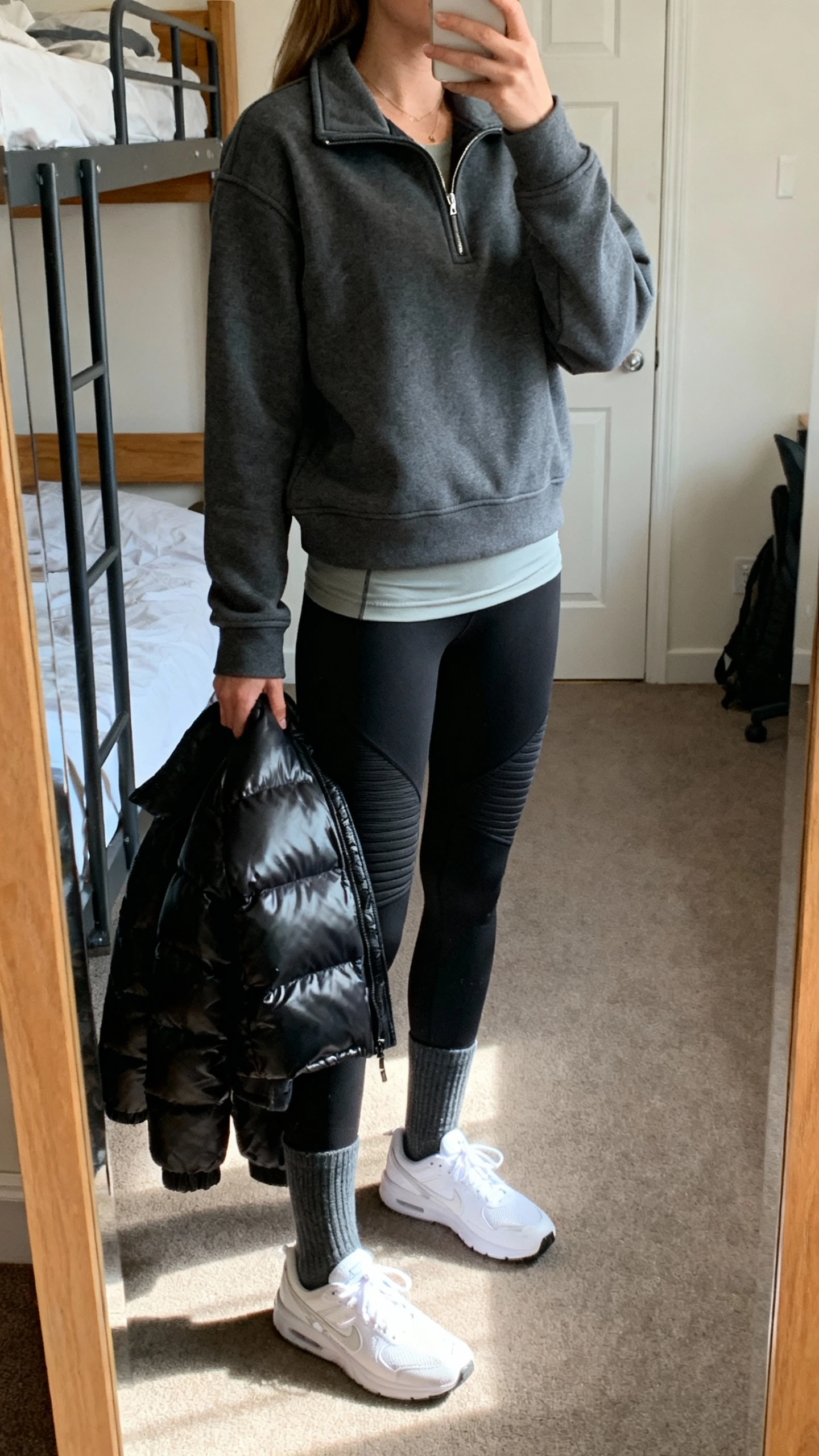 iPhone side-angle mirror selfie of a woman wearing a charcoal quarter-zip sweatshirt with structured collar, black thermal leggings, clean white supportive sneakers, long socks in a coordinating shade, a thin merino base layer, and a sleek black parka draped over arm, face not visible, dorm room full-length mirror, indoor morning light, iPhone photo quality.