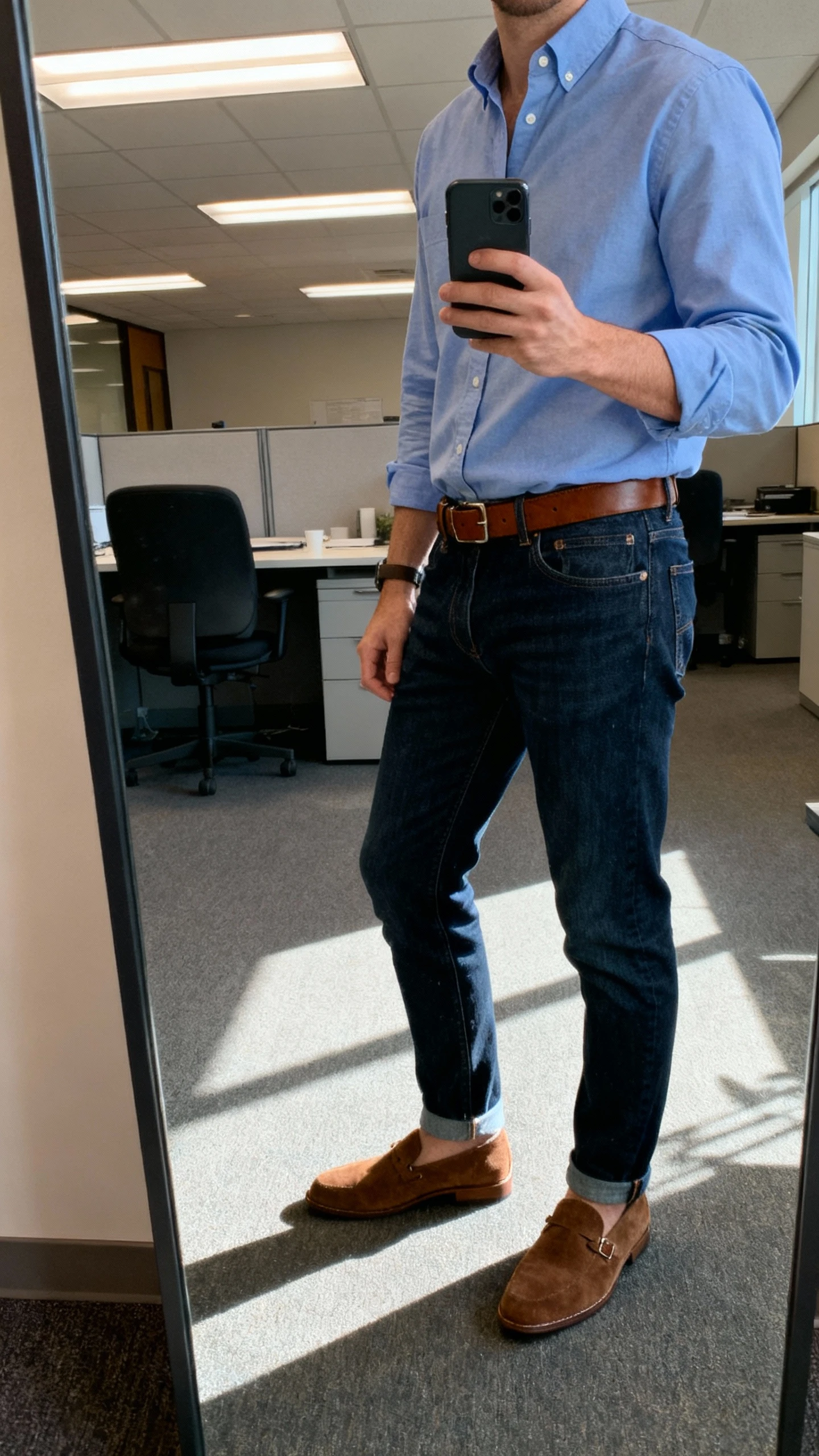 iPhone side-angle office mirror selfie of a man wearing a lightweight blue Oxford button-down with dark indigo jeans, brown leather belt and suede loafers, face not visible, fluorescent office lighting mixed with daylight, casual iPhone photo.