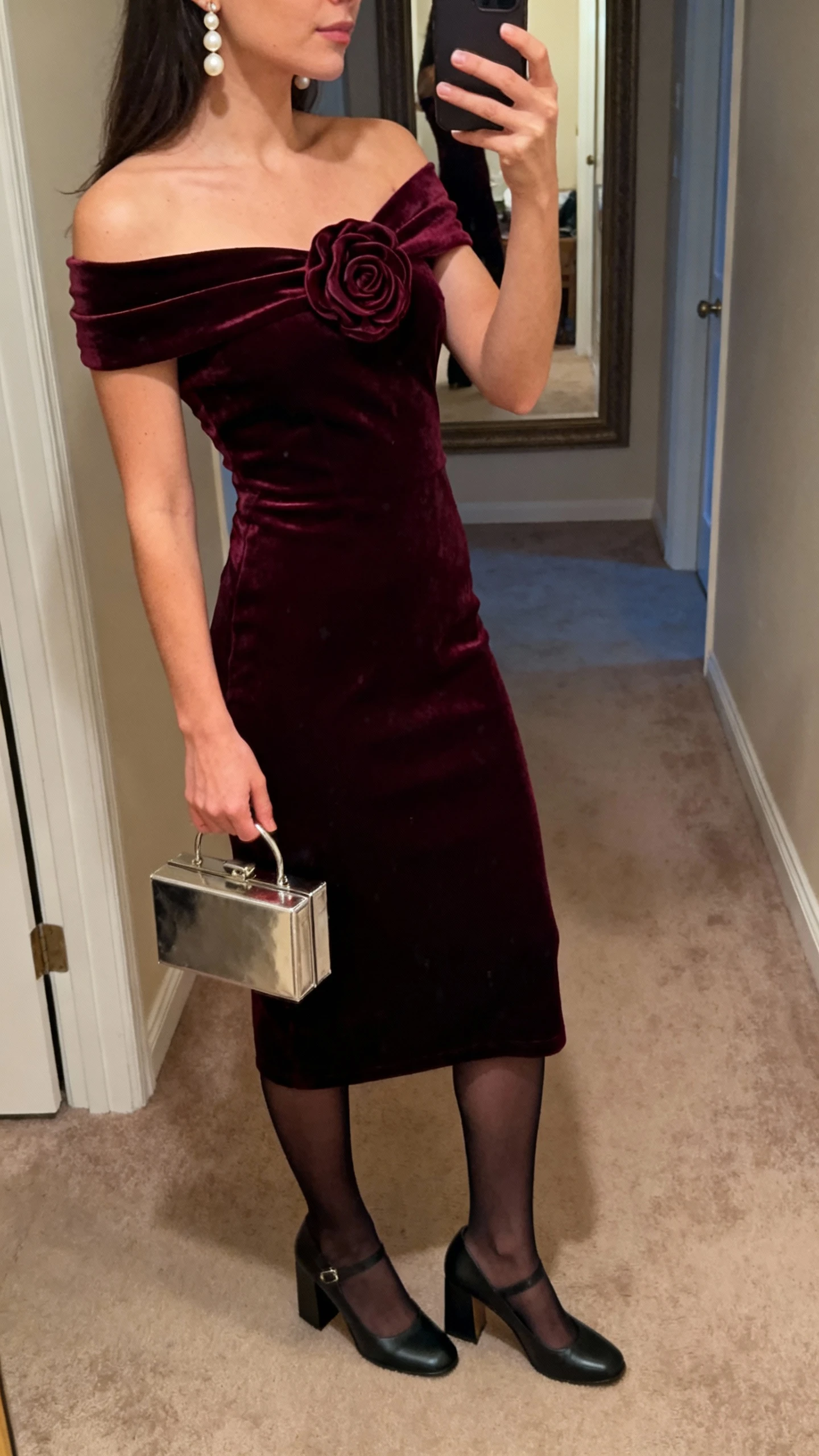 iPhone side-angle selfie cropped at shoulders showing a woman wearing an off-the-shoulder velvet midi dress in deep wine with a statement rosette, sheer black tights, block-heel Mary Janes, metallic mini box clutch, and pearl drop earrings; face not visible, hallway mirror, warm evening light, casual iPhone photo.