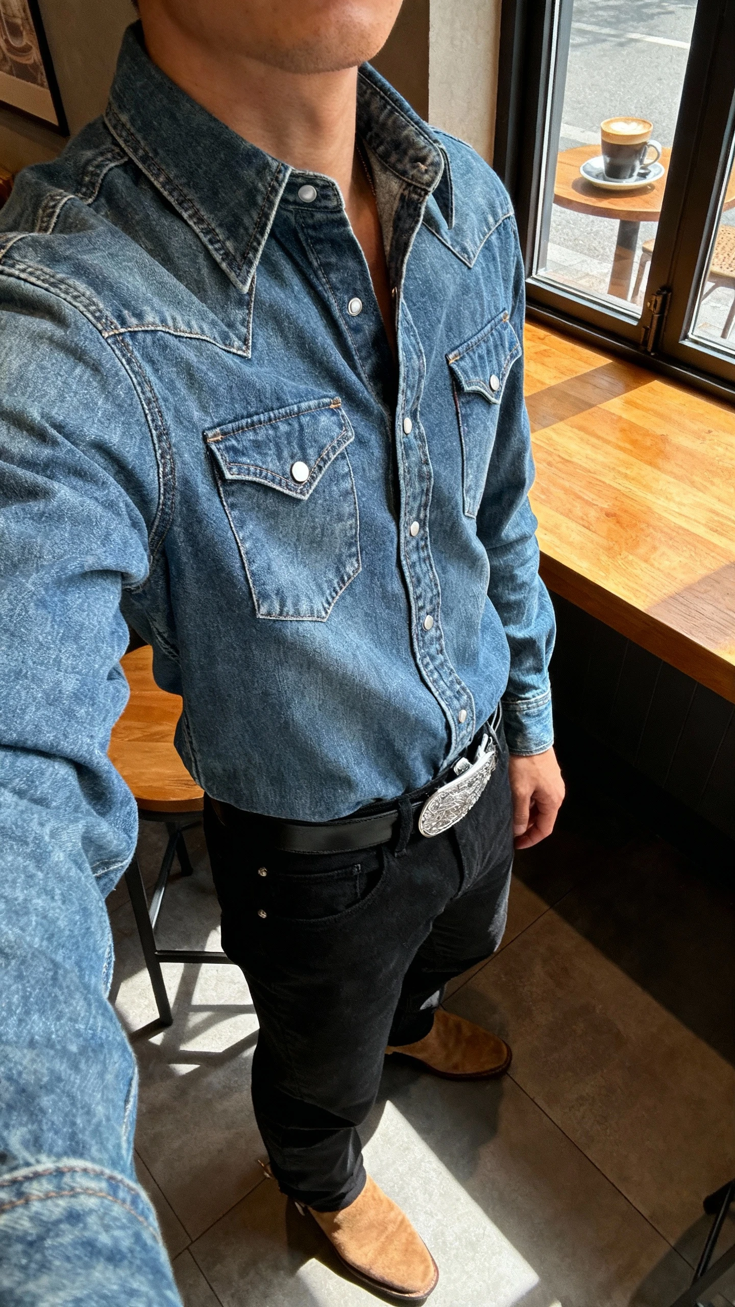 iPhone side-angle selfie of a man in a snap-front Western denim shirt tucked into black jeans with a silver-buckle belt and suede boots, face not visible, coffee shop window light on a wooden table backdrop, natural iPhone quality.
