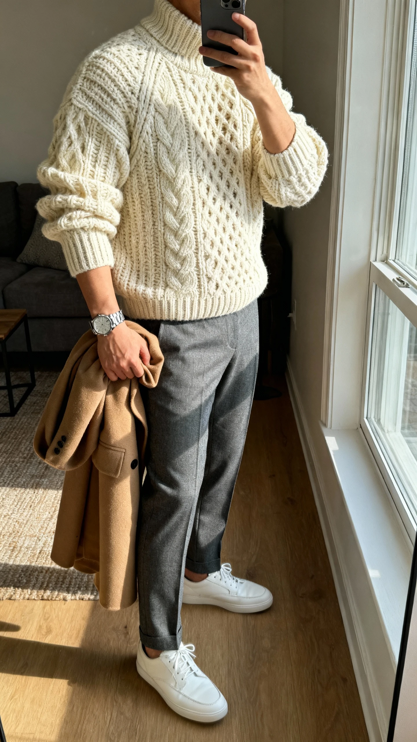 iPhone side-angle selfie of a man wearing a chunky cream fisherman knit sweater, tailored grey wool trousers, white leather sneakers, camel overcoat draped over arm, stainless watch, face not visible, living room by window, soft morning light, natural iPhone quality