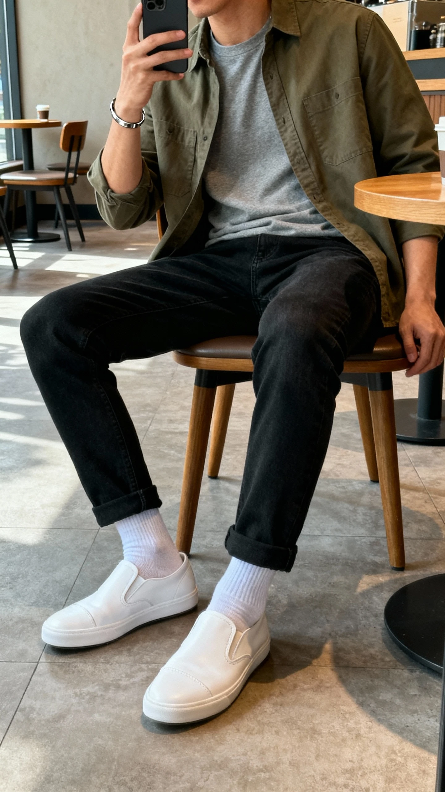 iPhone side-angle selfie of a man wearing white low-profile leather sneakers, black tapered jeans, an olive cotton-twill overshirt over a gray tee, invisible ankle socks, and a slim steel bracelet; casual coffee shop seating, natural daylight, face not visible, natural iPhone photo.