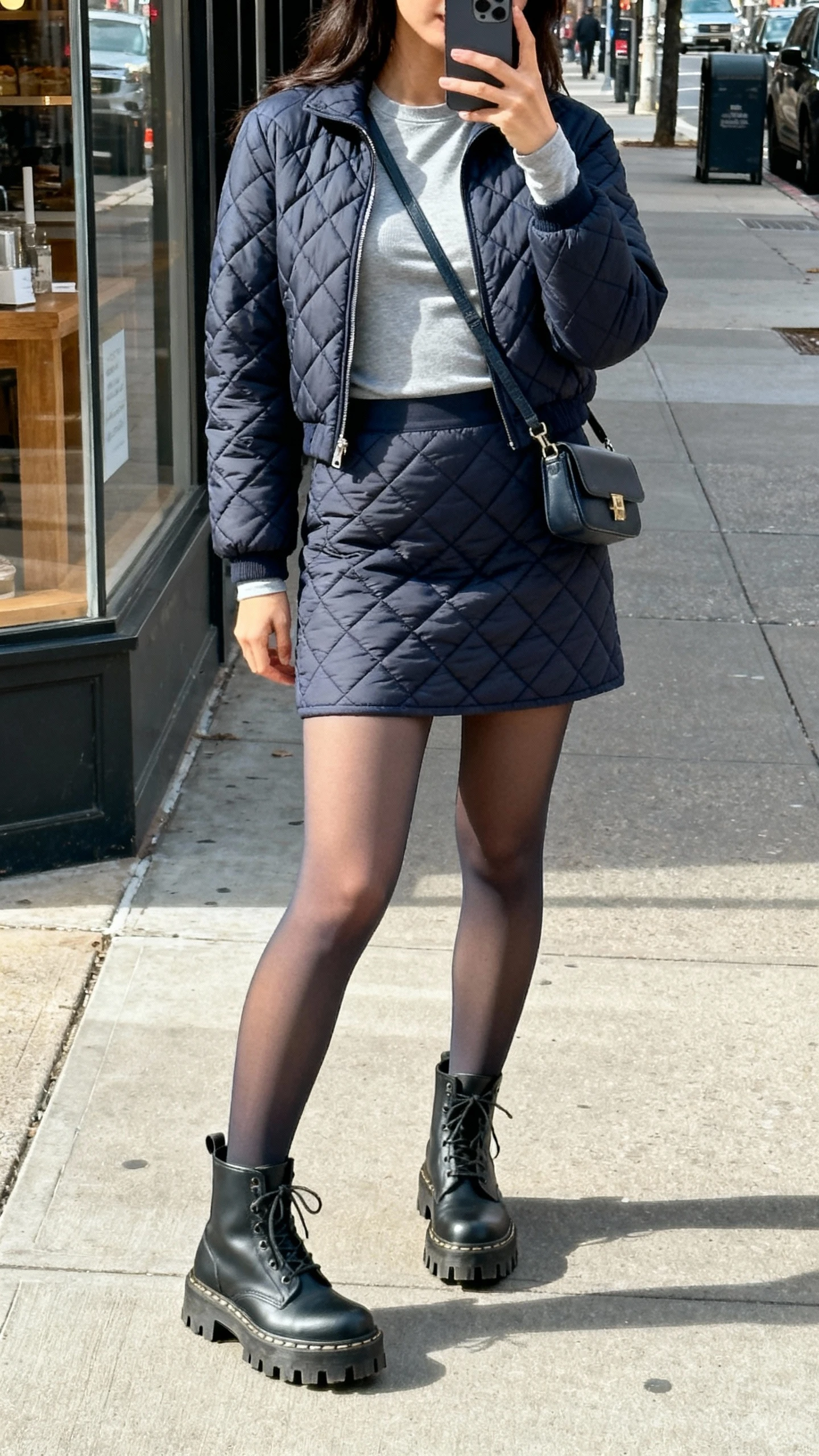 iPhone side-angle selfie of a woman in a quilted skirt set (quilted mini skirt and matching jacket), fitted long-sleeve tee underneath, opaque tights, lug-sole ankle boots, small crossbody, face not visible, city sidewalk near storefront, natural daylight, casual iPhone photo.
