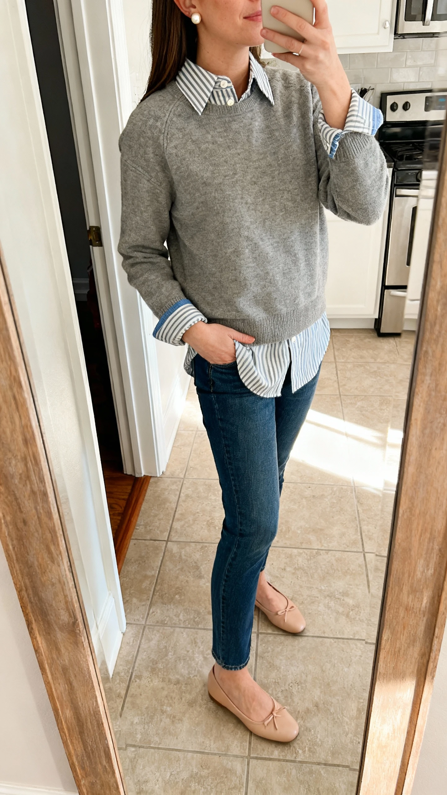 iPhone side-angle selfie of a woman wearing a crewneck sweater over a striped button-down with the collar, hem, and cuffs showing, straight-leg jeans, ballet flats, pearl studs, face not visible, kitchen doorway mirror, morning light, natural iPhone photo.