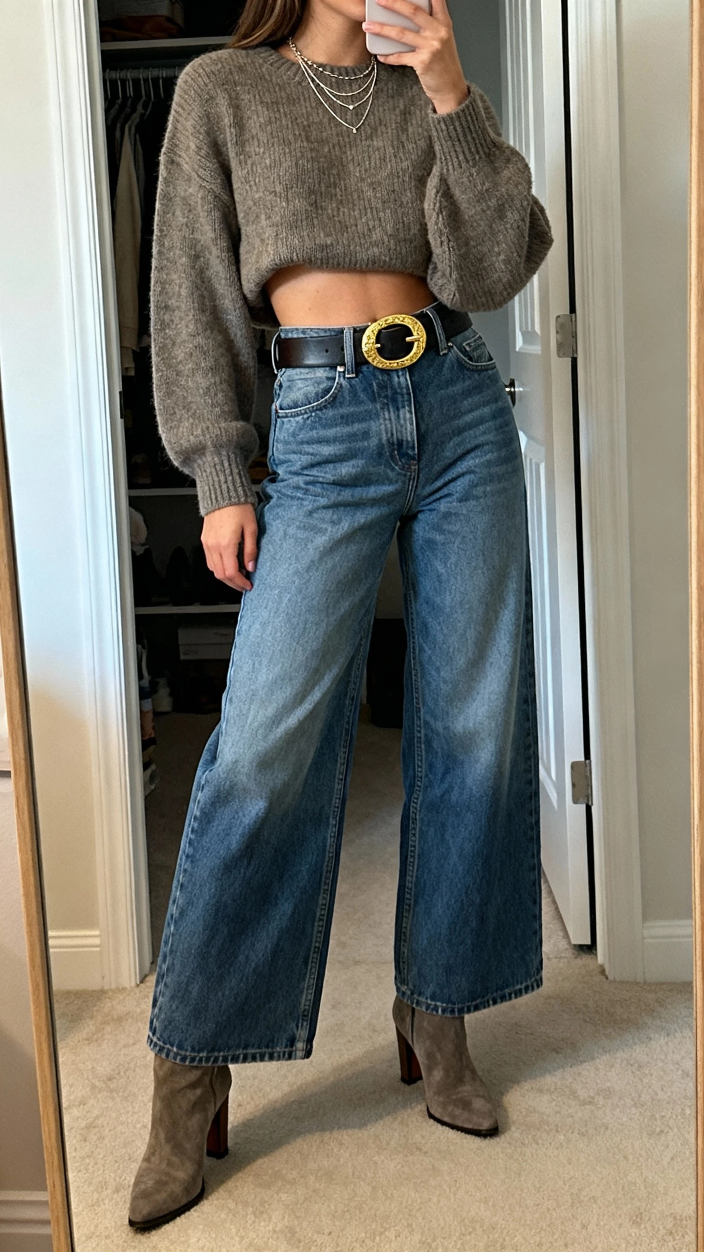 iPhone side-angle selfie of a woman wearing high-rise wide-leg jeans, a cropped sweater, a bold statement belt with a large buckle, heeled ankle boots, and delicate layered necklaces, face not visible, cropped at neck, closet mirror, indoor lighting, iPhone photo quality.