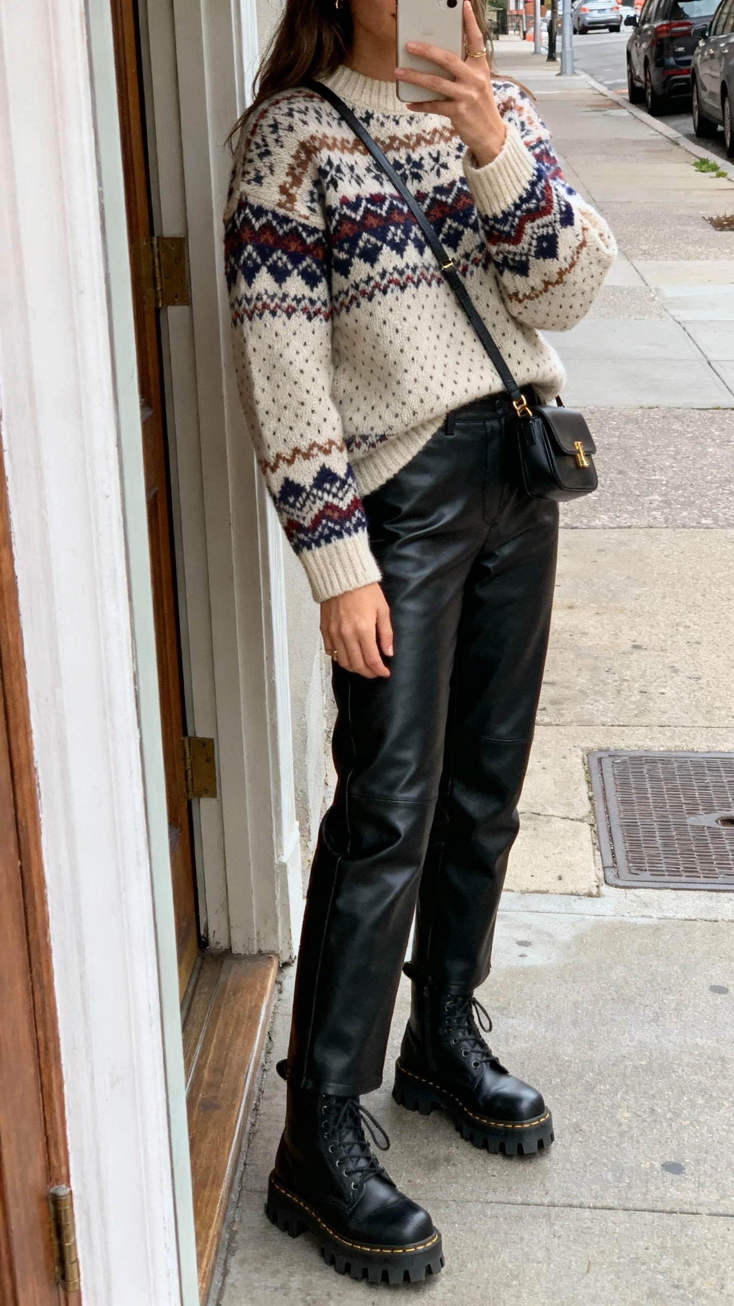 iPhone side-angle selfie showing a woman in a Fair Isle sweater with black leather pants, lug-sole boots, small crossbody bag, face not visible, city street doorway, soft daylight, natural iPhone photo.