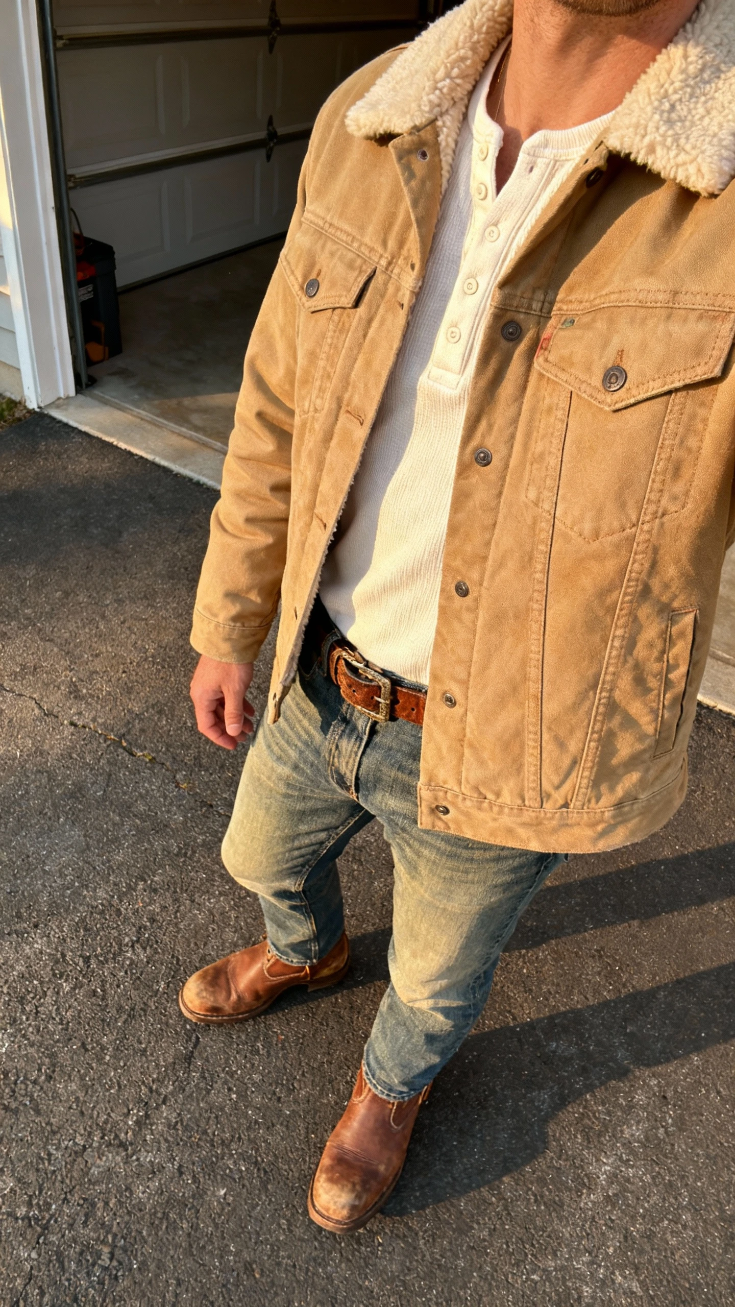 iPhone side-profile selfie of a man wearing a tan shearling-collar trucker jacket over an off-white henley, medium-wash denim, brown leather work boots, and a rugged leather belt; garage/driveway setting, late afternoon light, cropped at shoulders, face not visible, natural iPhone photo.