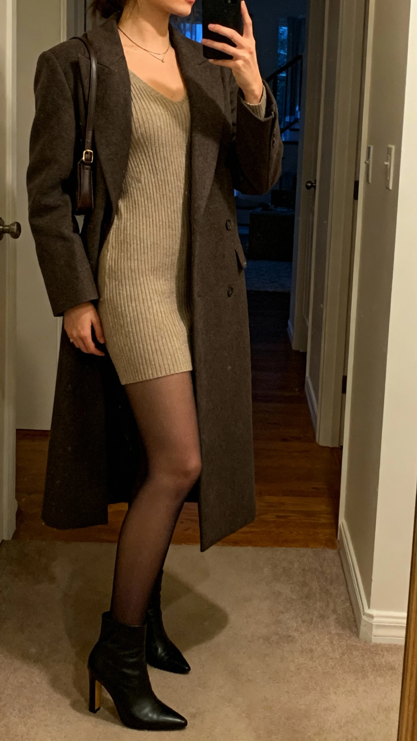 iPhone side-view selfie of a woman in a ribbed knit dress with sheer tights and a long tailored coat, heeled ankle boots, minimal necklace, small shoulder bag, face not visible, apartment hallway mirror, evening indoor light, natural iPhone aesthetic.