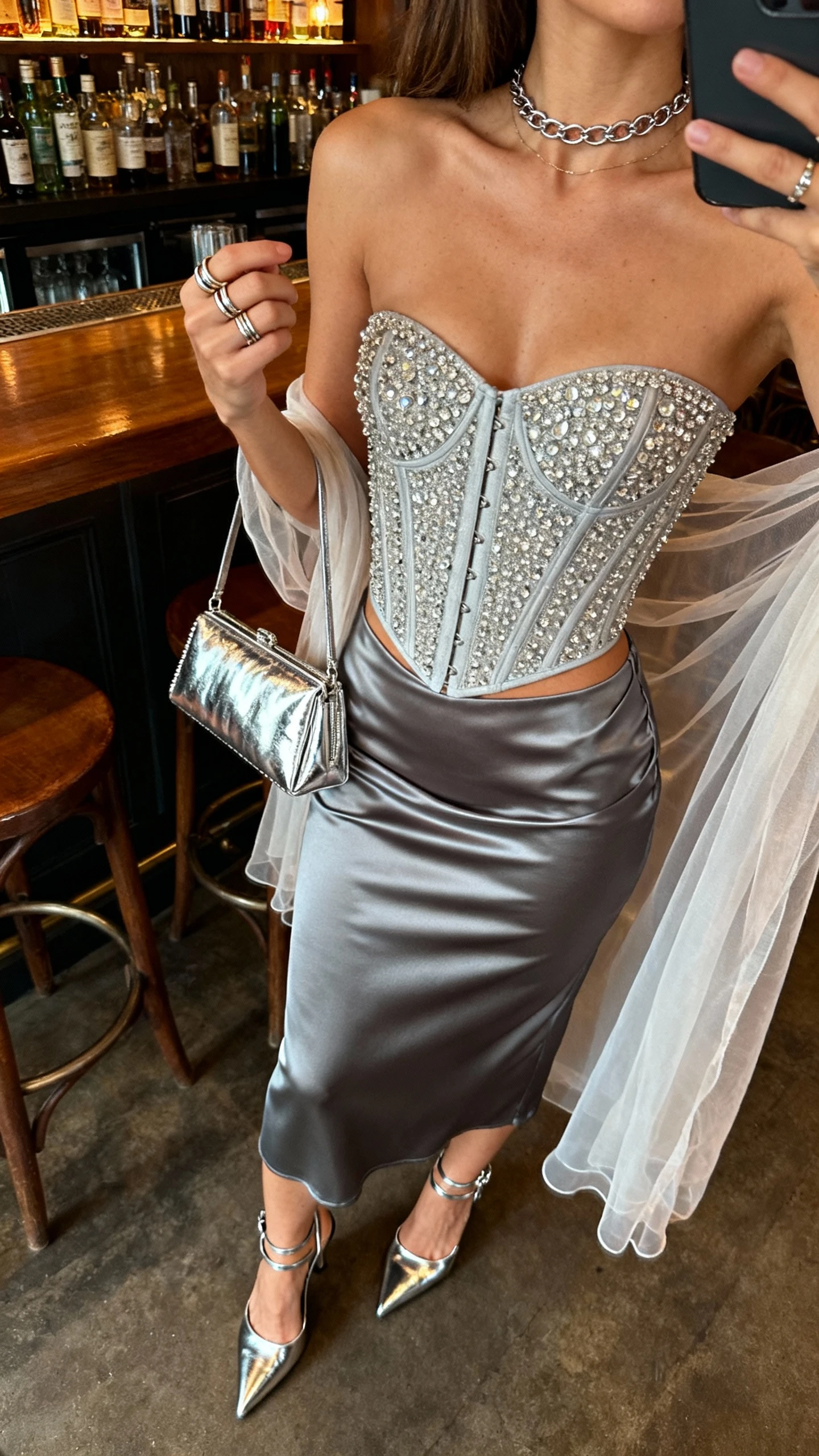 iPhone sitting-at-bar selfie cropped at neck of a woman wearing a rhinestone corset top, gunmetal satin bias-cut midi skirt, metallic ankle-strap heels, shiny baguette bag, delicate chain choker, stacking rings, and a sheer shawl; face not visible, cozy bar lighting, natural iPhone aesthetic.
