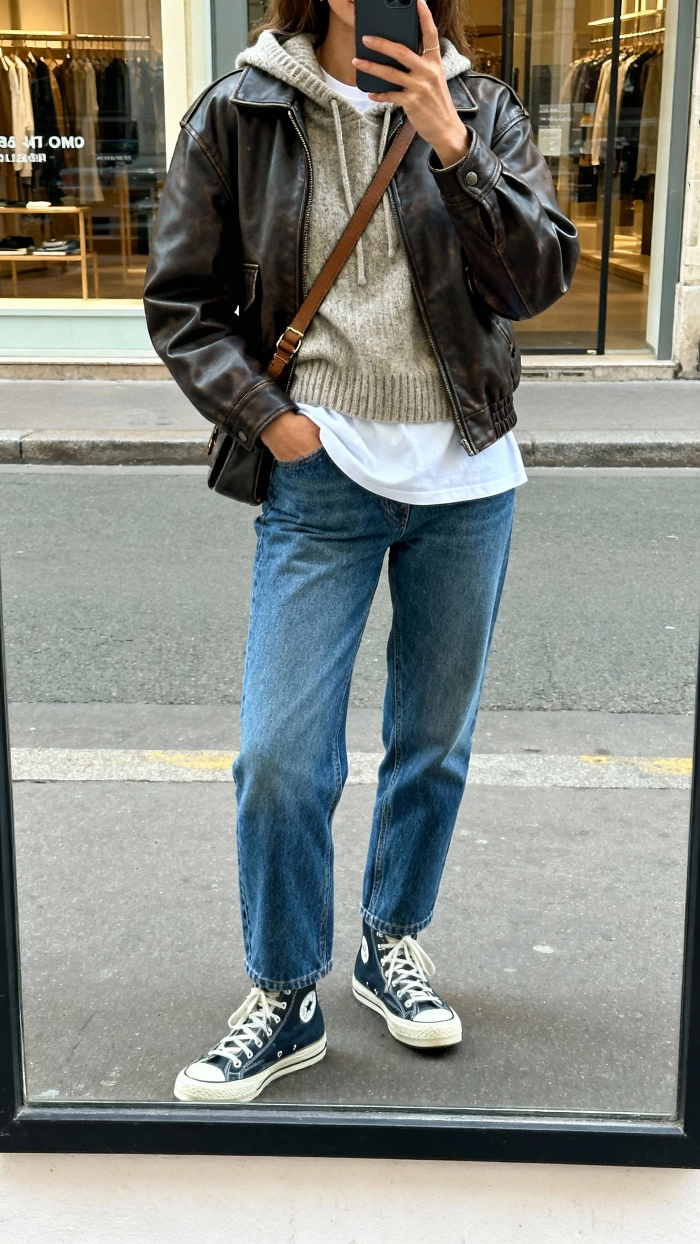 iPhone street-mirror selfie of a woman wearing a leather jacket layered over a knit hoodie and a white tee, straight blue jeans, high-top sneakers, crossbody bag, face not visible, natural daylight, urban storefront reflection, iPhone photo quality.