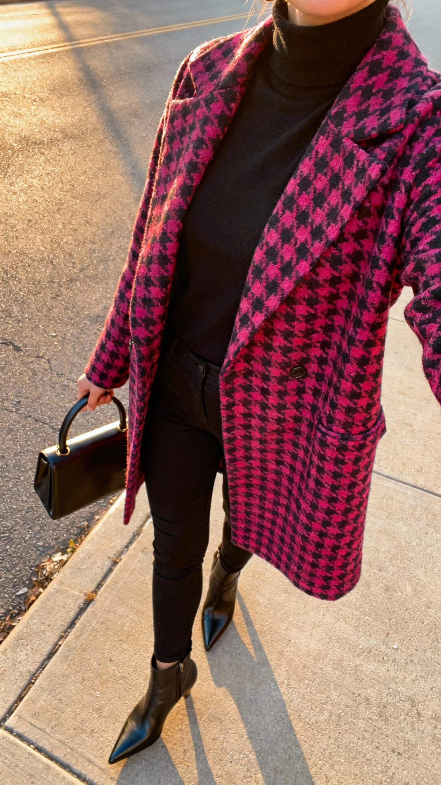 iPhone street-side selfie, cropped at neck, of a woman in a sleek all-black base (black turtleneck and slim black pants) topped with a bold statement coat (vibrant pattern), pointed ankle boots, structured top-handle bag, face not visible, golden-hour light, casual iPhone aesthetic